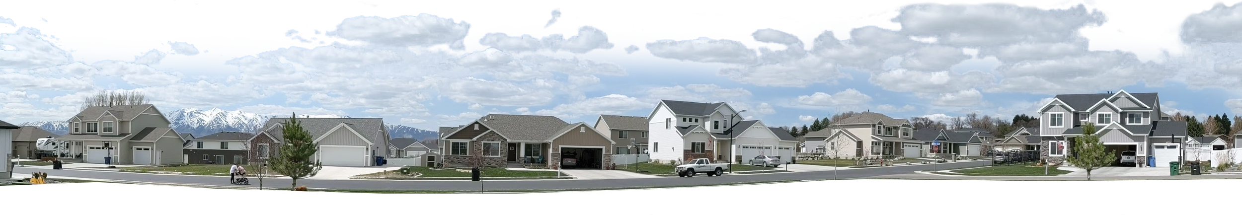 A panoramic view of a suburban neighborhood with multiple modern houses, some with attached garages, on a sunny day with a partly cloudy sky, and snow-capped mountains in the background.