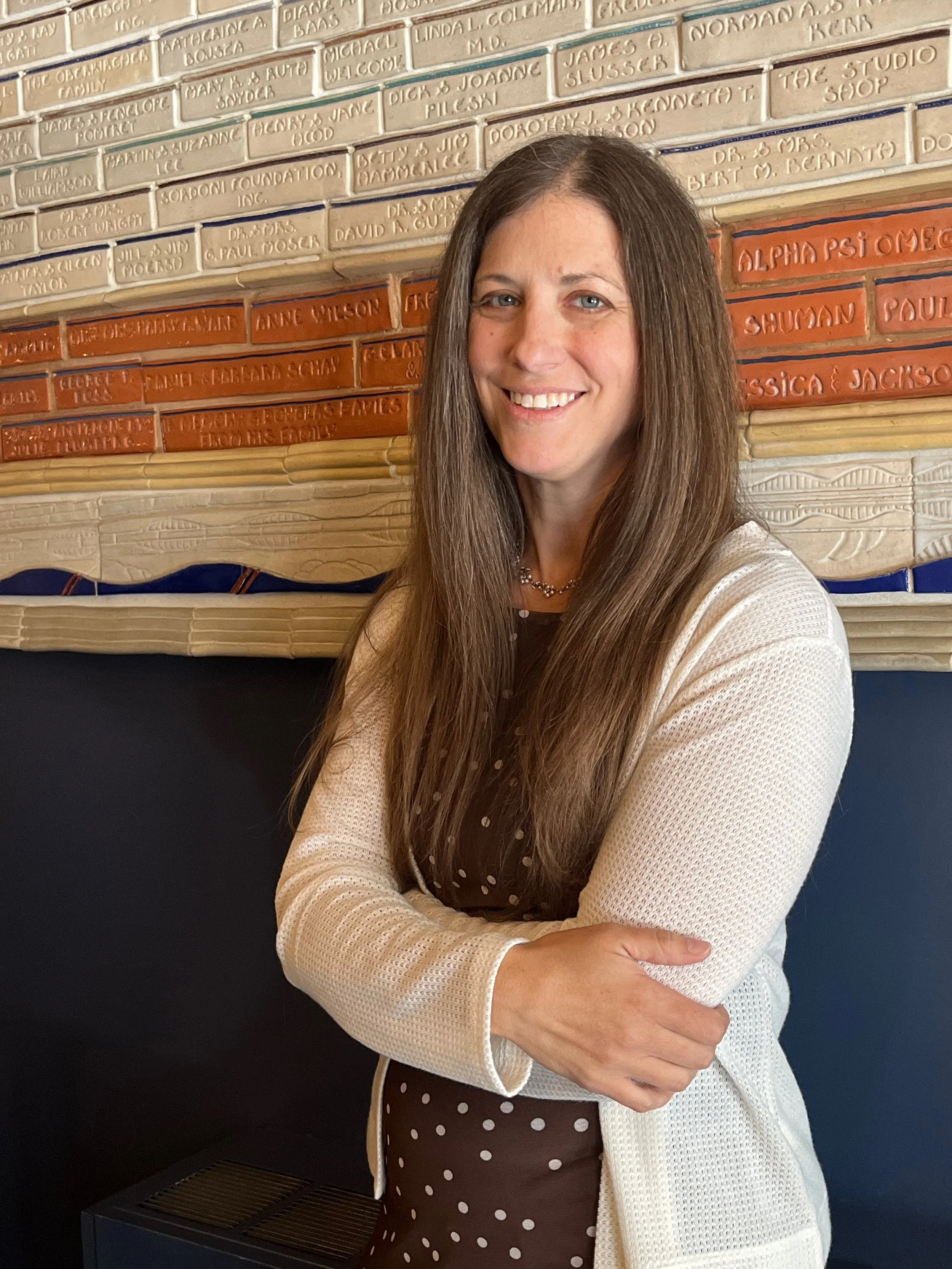 A woman with long brown hair and blue eyes smiling in front of a brick wall in the BTE lobby. .