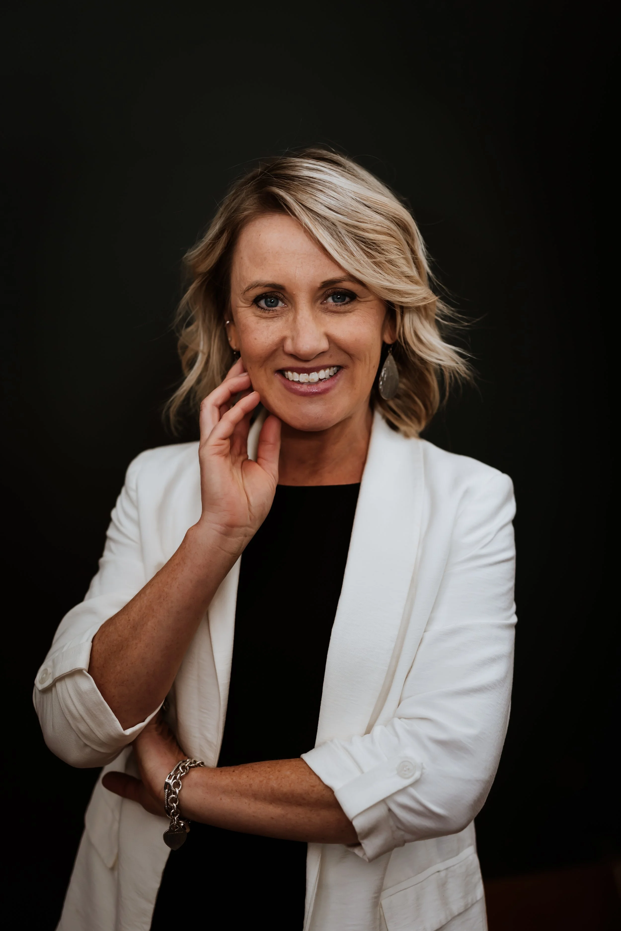 Real Estate Agent Samantha Capita stands smiling at the camera, her left hand resting on her chin, against a black backdrop. She is a white woman with balayage blonde hair wearing a black top, white blazer, and dangling earrings. 