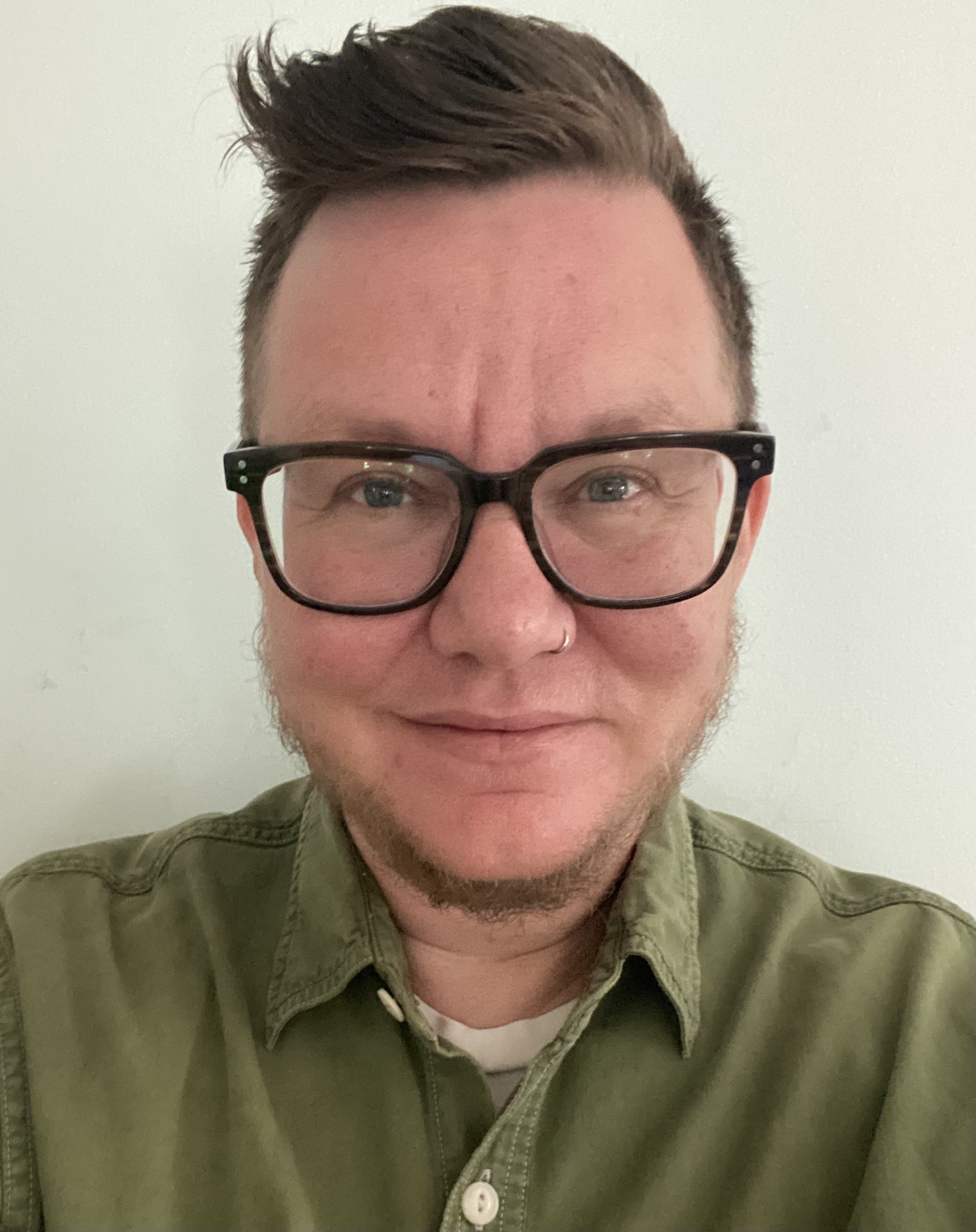 Dr. Llewelyn “Lew” Lewis wears a green tailored shirt, has a chinstrap beard, a nose-ring, and square glasses. Dr. Lewis stands in front of a mint green wall.