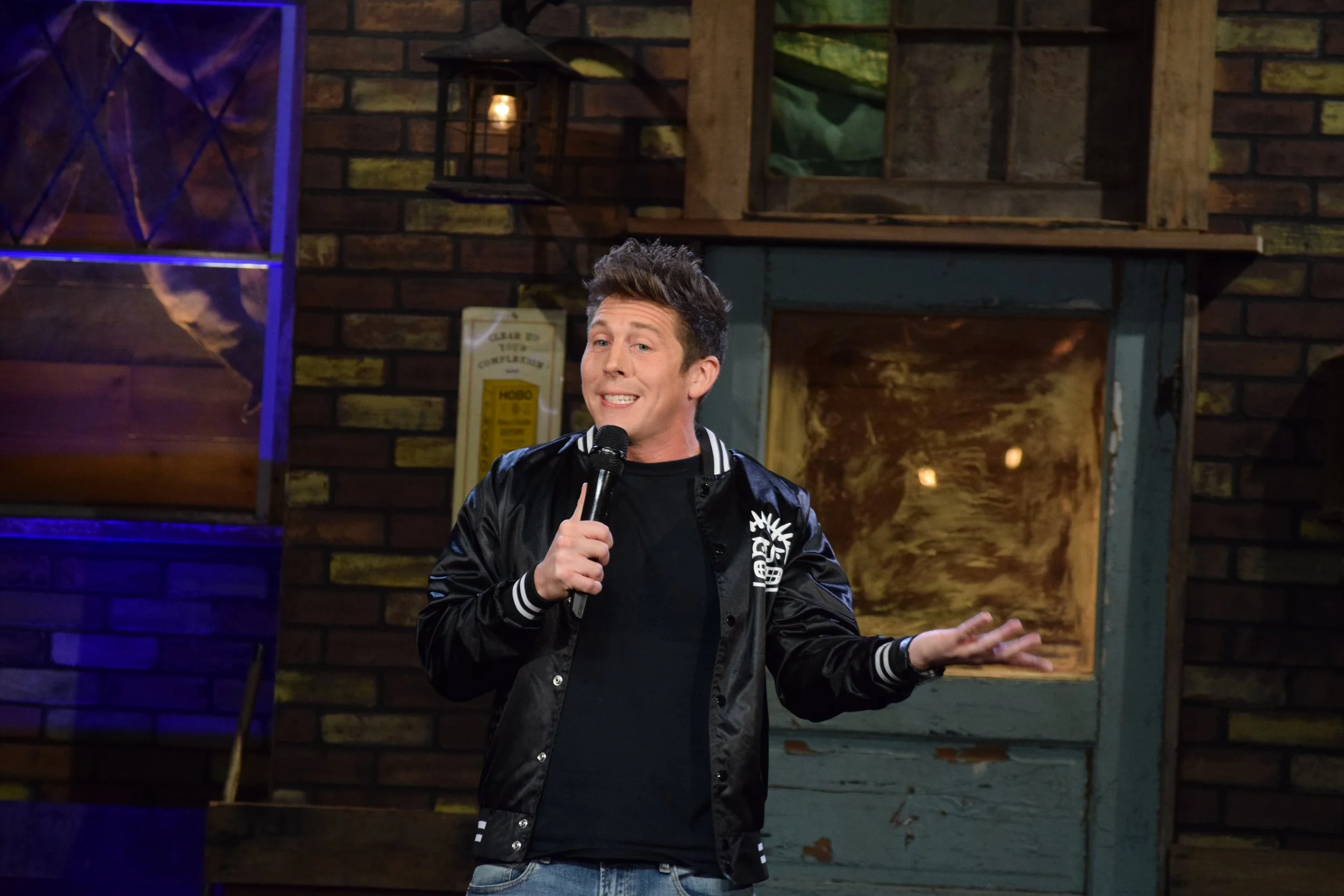 Lee Hardin, a white man with spiky brown hair in a sports coat, holds a microphone to his face.