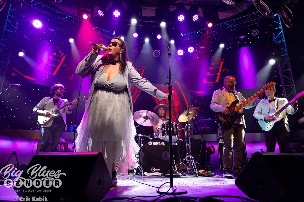 A woman in a grey mini dress, with ripples of ethereal gauze going past the skirt's hem to the floor. Clad in sunglass, she sings with her band of three string instrument players and a drummer on a purple-lighted stage.