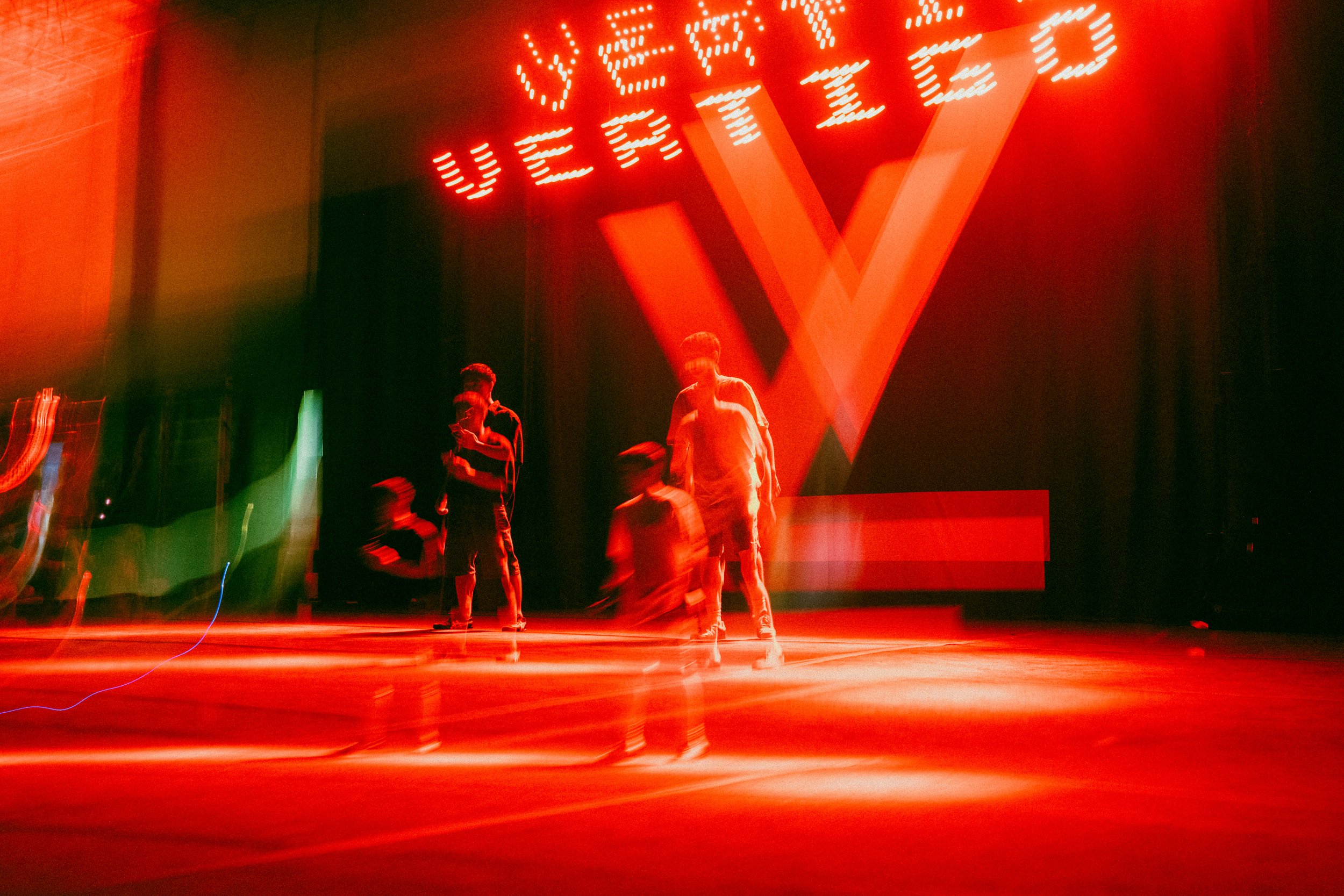 Red neon sign that reads 'VETEZ' in a dark room with people’s silhouettes and a large V-shaped logo projected on the wall.