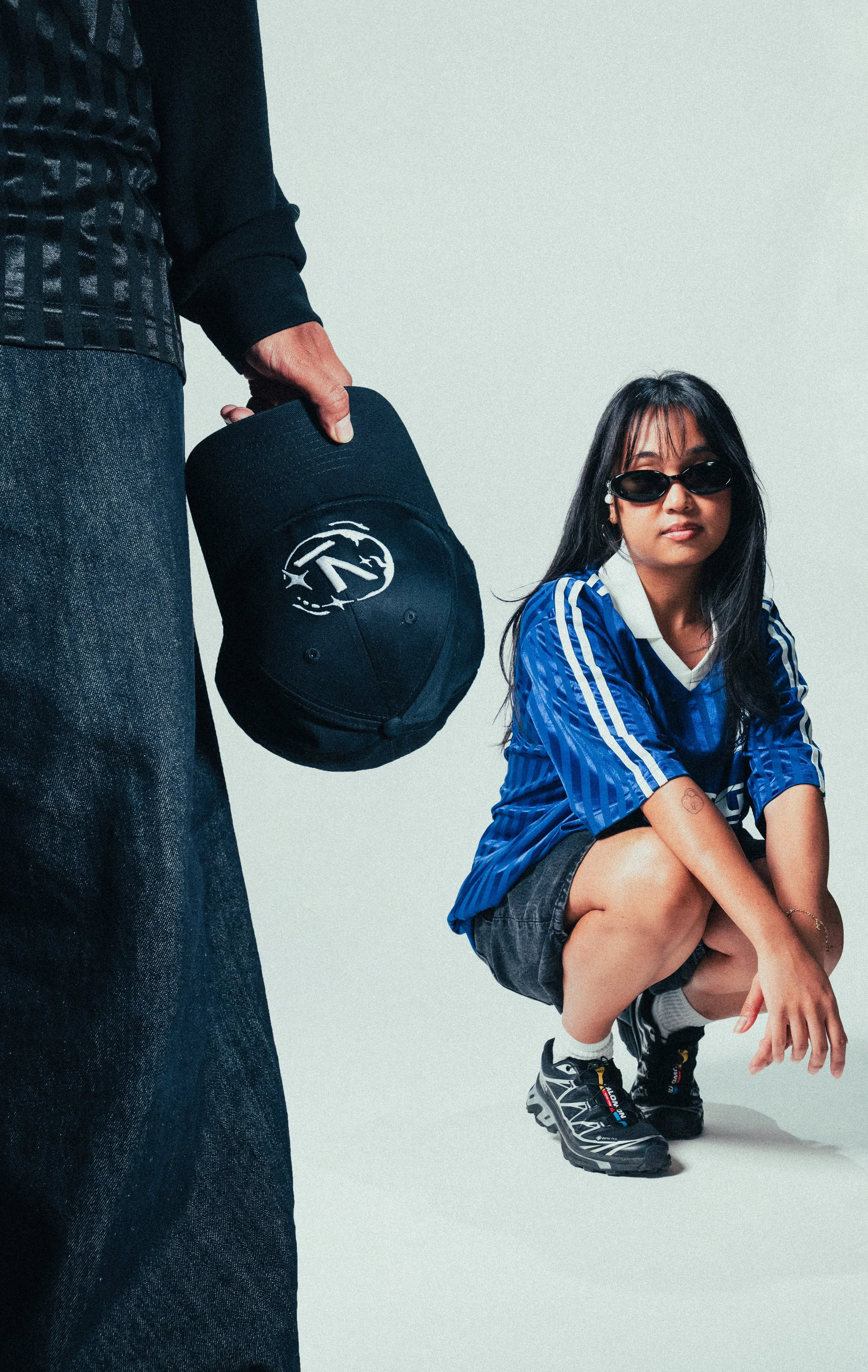 A woman wearing sunglasses squatting with her arms resting on her knees, wearing a blue sports jersey, denim shorts, and sneakers. A person standing nearby holds a black cap with a logo.