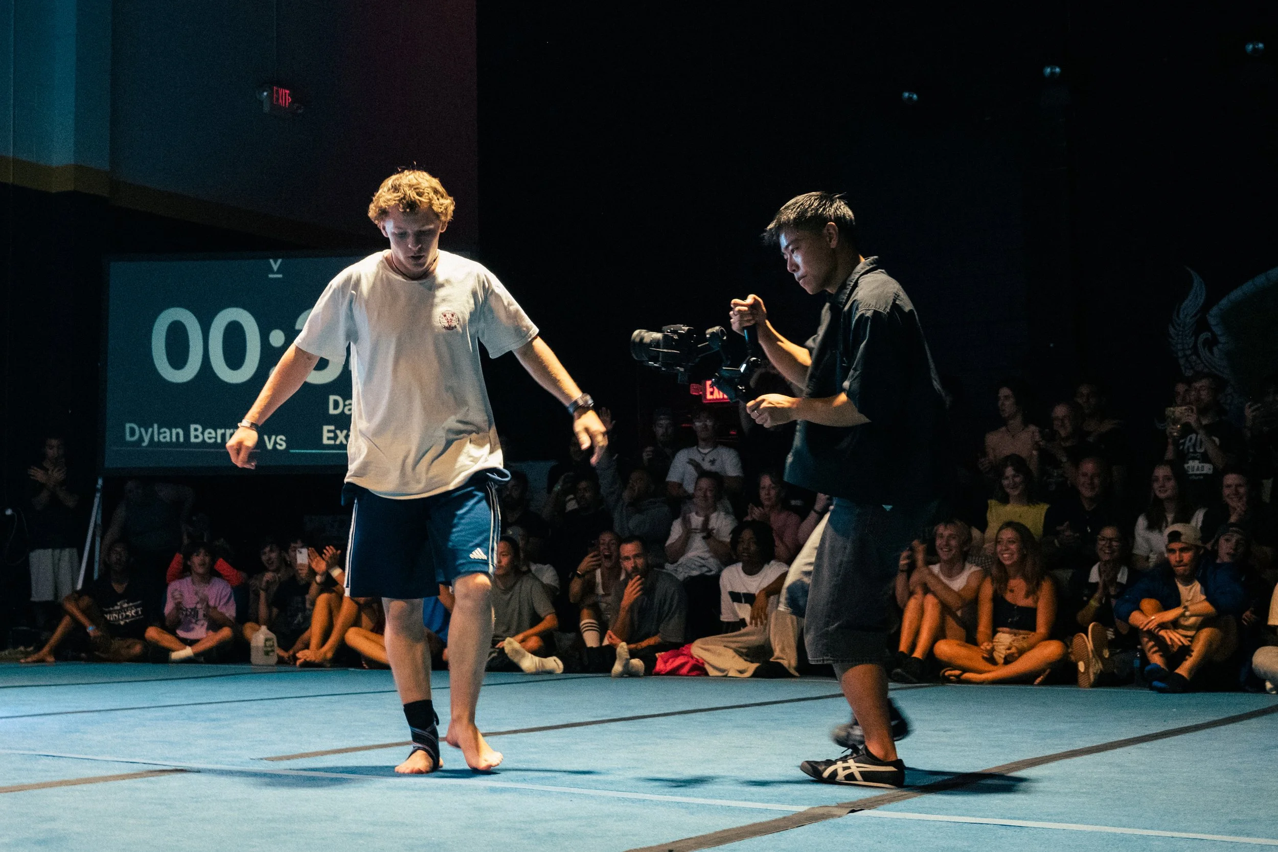 Two young men are performing a dance on a stage in front of a live audience. The person on the left has curly hair, is wearing a white t-shirt, blue shorts, and ankle socks, and appears to be in movement. The person on the right has straight hair, is wearing a dark shirt and pants, and is holding an electronic device. Behind them, there is a large digital timer and a crowd of spectators sitting and watching.