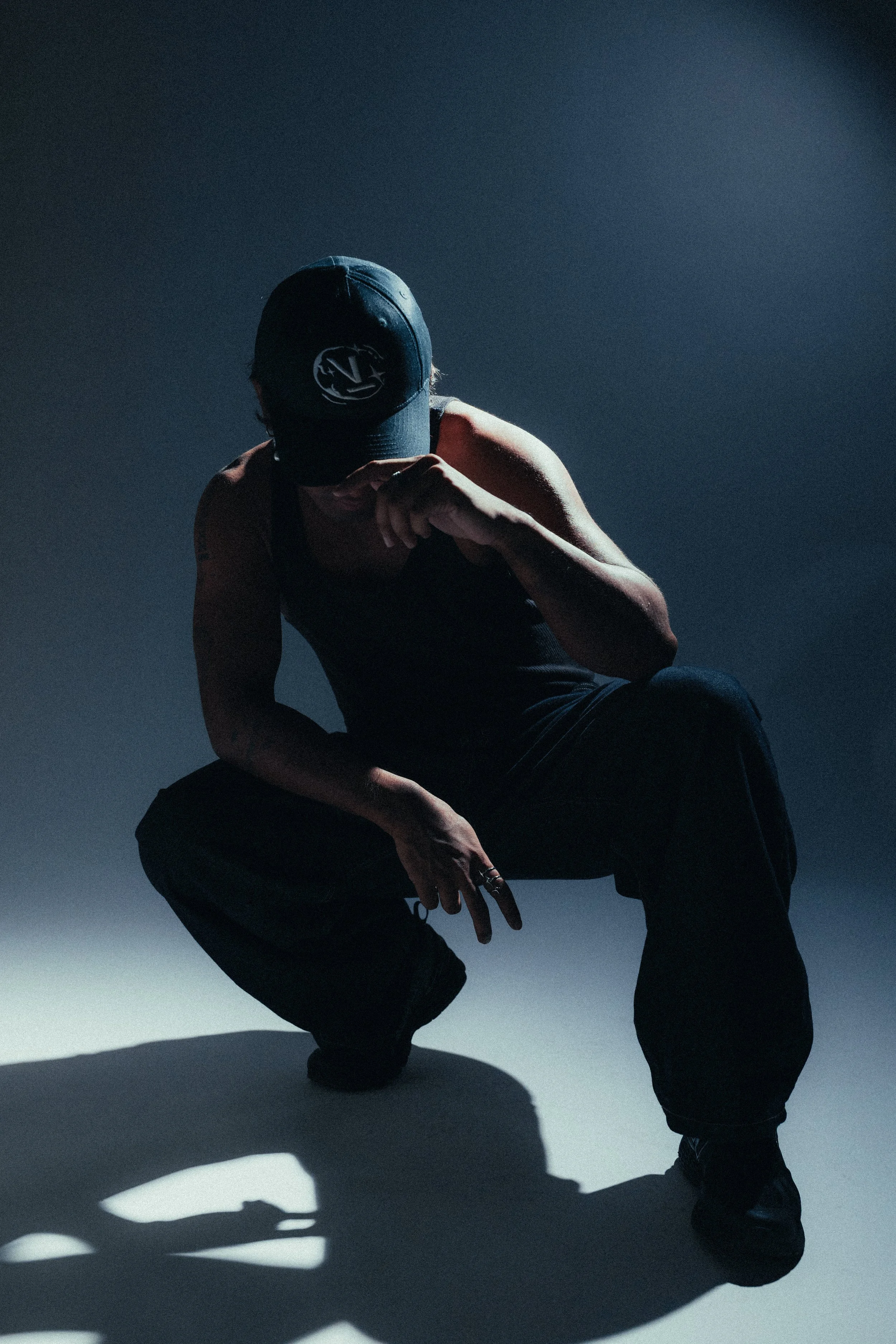 Person crouching down, wearing a black baseball cap with a logo, black pants, and black shoes, casting shadows on the floor.