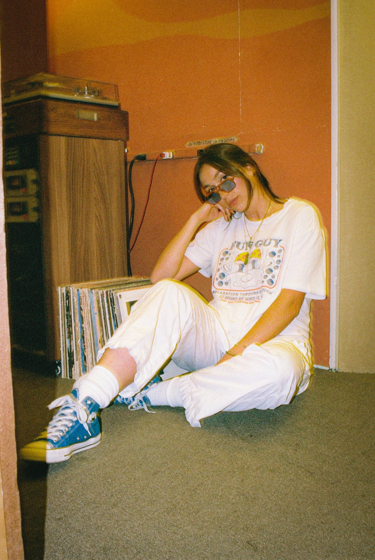 A young woman with long brown hair leaning forward outdoors in a green park, wearing a white t-shirt with green text, surrounded by trees and grass.