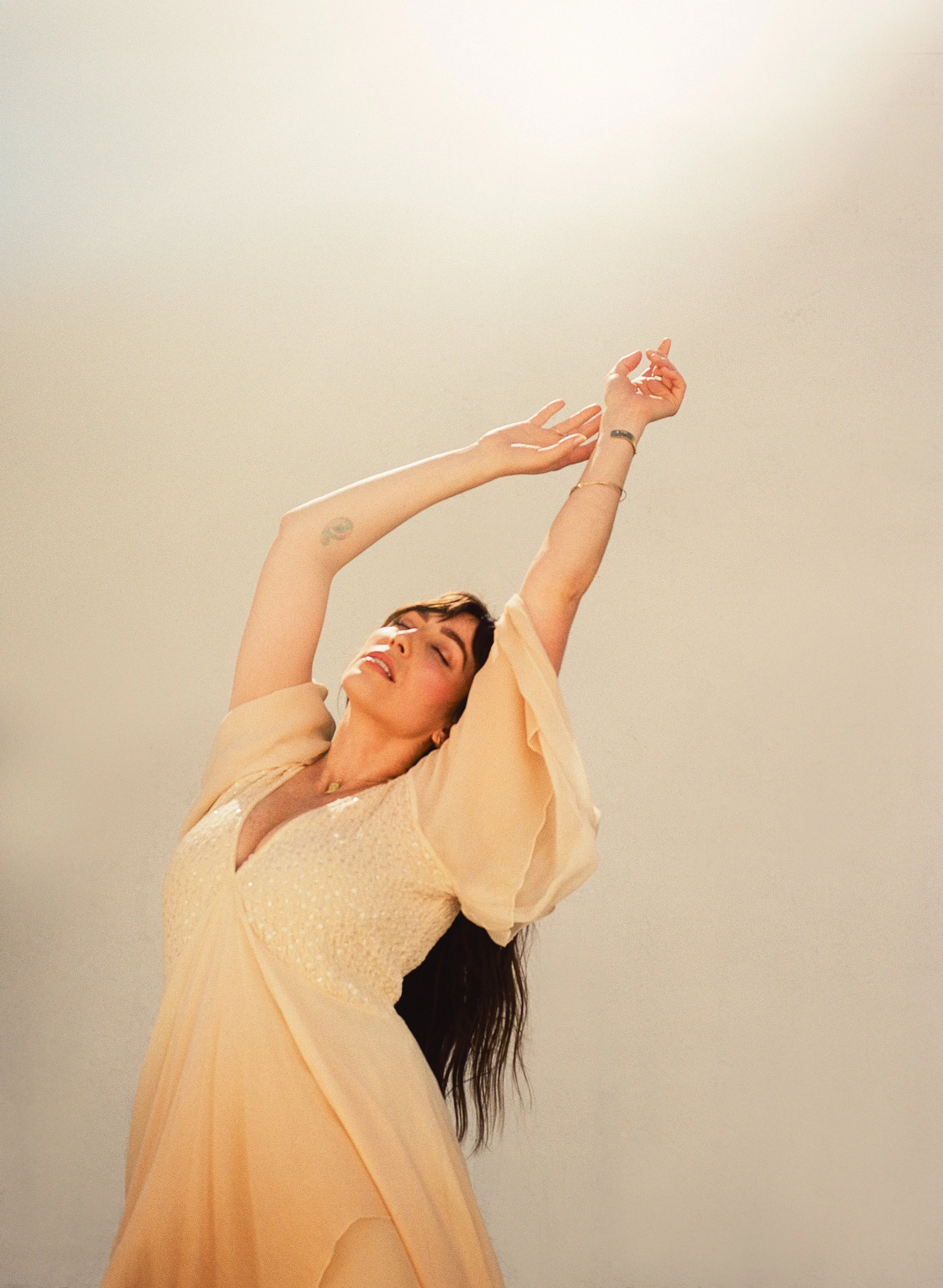 Singer-Songwriter  Abigail Rose with long dark hair dancing in a flowing peach-colored dress, arms raised, eyes closed, against a plain light background