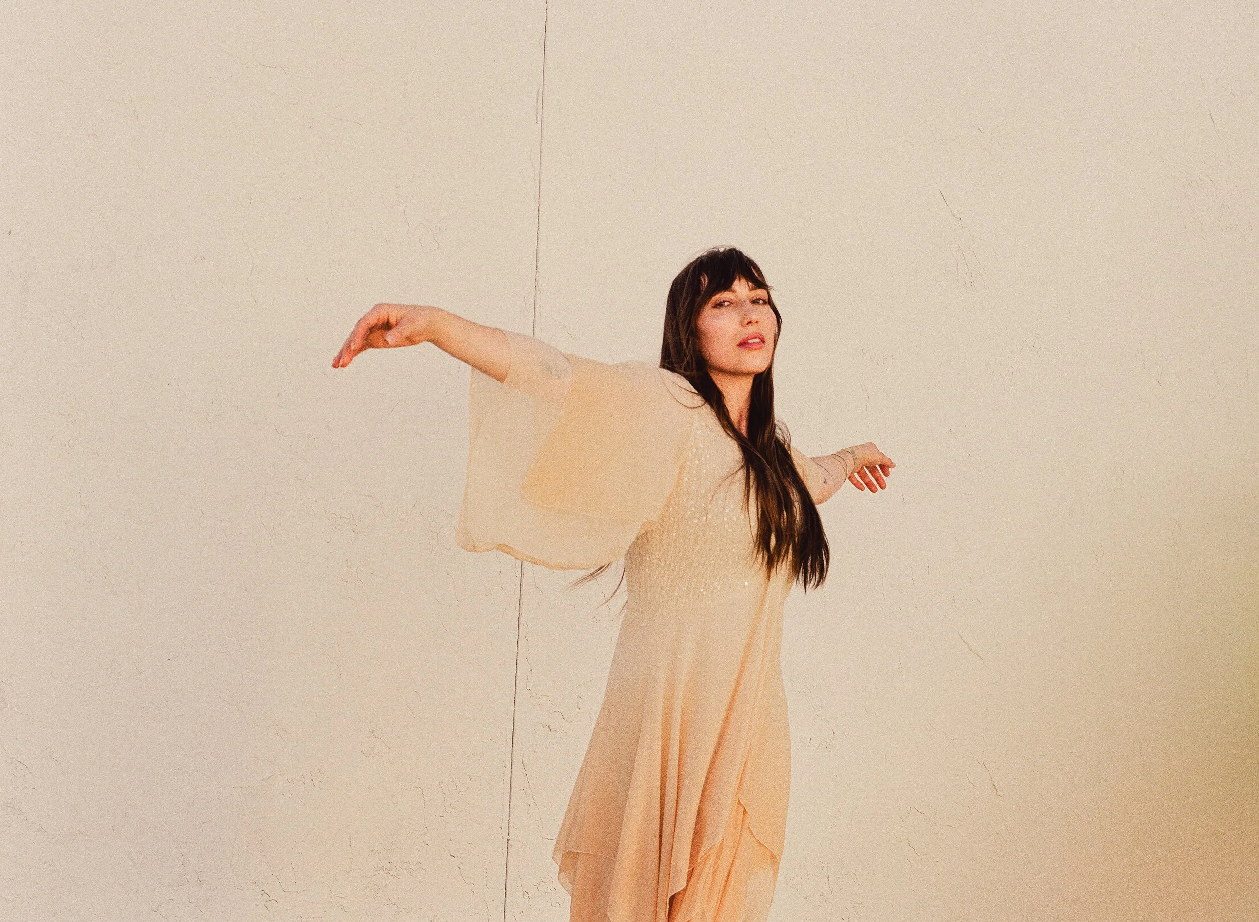 Singer-Songwriter Abigail Rose with long dark hair dancing or posing with her arms spread wide in front of a plain light-colored wall.