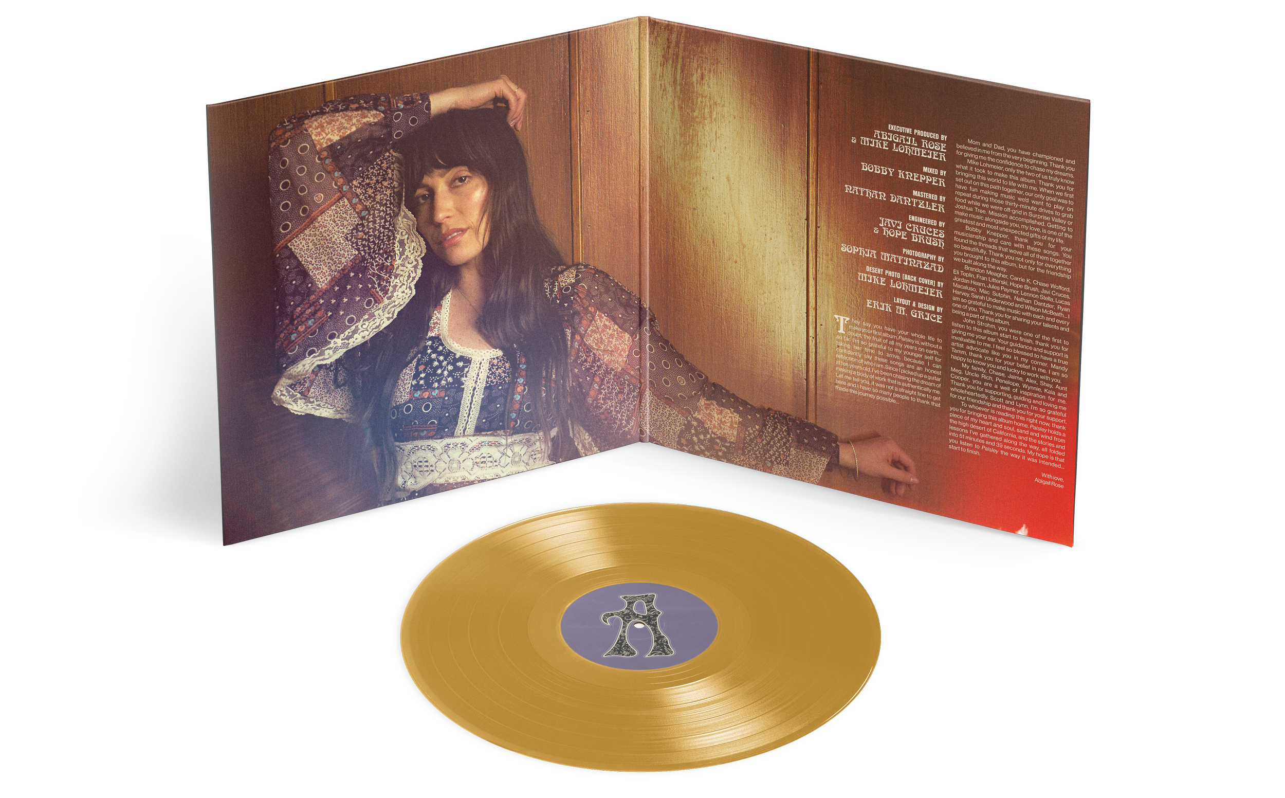 A gold vinyl record with a purple label featuring a metallic number 5, in front of an open album cover showing a woman with long dark hair wearing a patterned dress, resting her head on one hand with a background of wood paneling and credits.