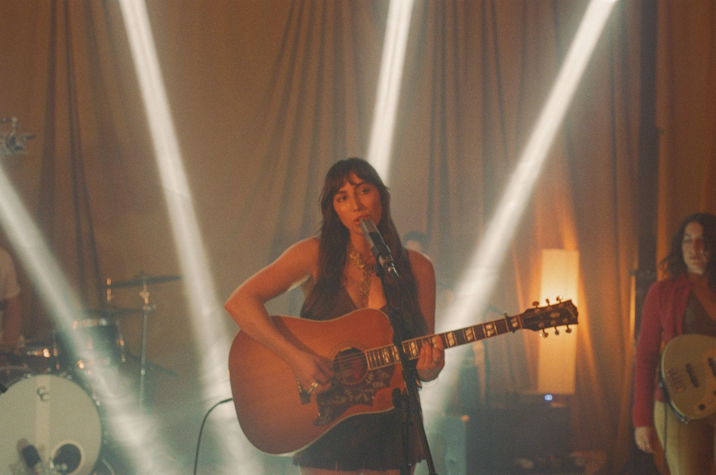 Singer-Songwriter  Abigail Ros with long dark hair and a floral top is playing an acoustic guitar and singing into a microphone. She is on a dimly lit stage with vertical beams of light behind her, and a woman in a red dress is standing to the right.