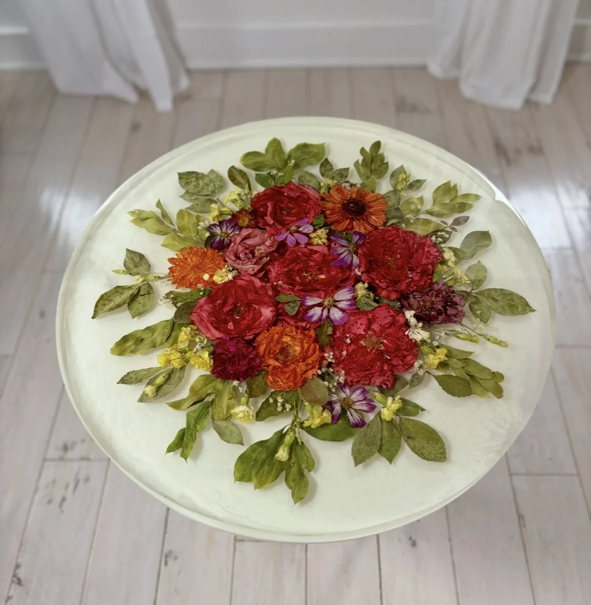 Top view of a white round resin table with a floral preservation  arrangement of red, orange, purple, and yellow flowers and green leaves on a white surface. Wedding bouquet resin table.
