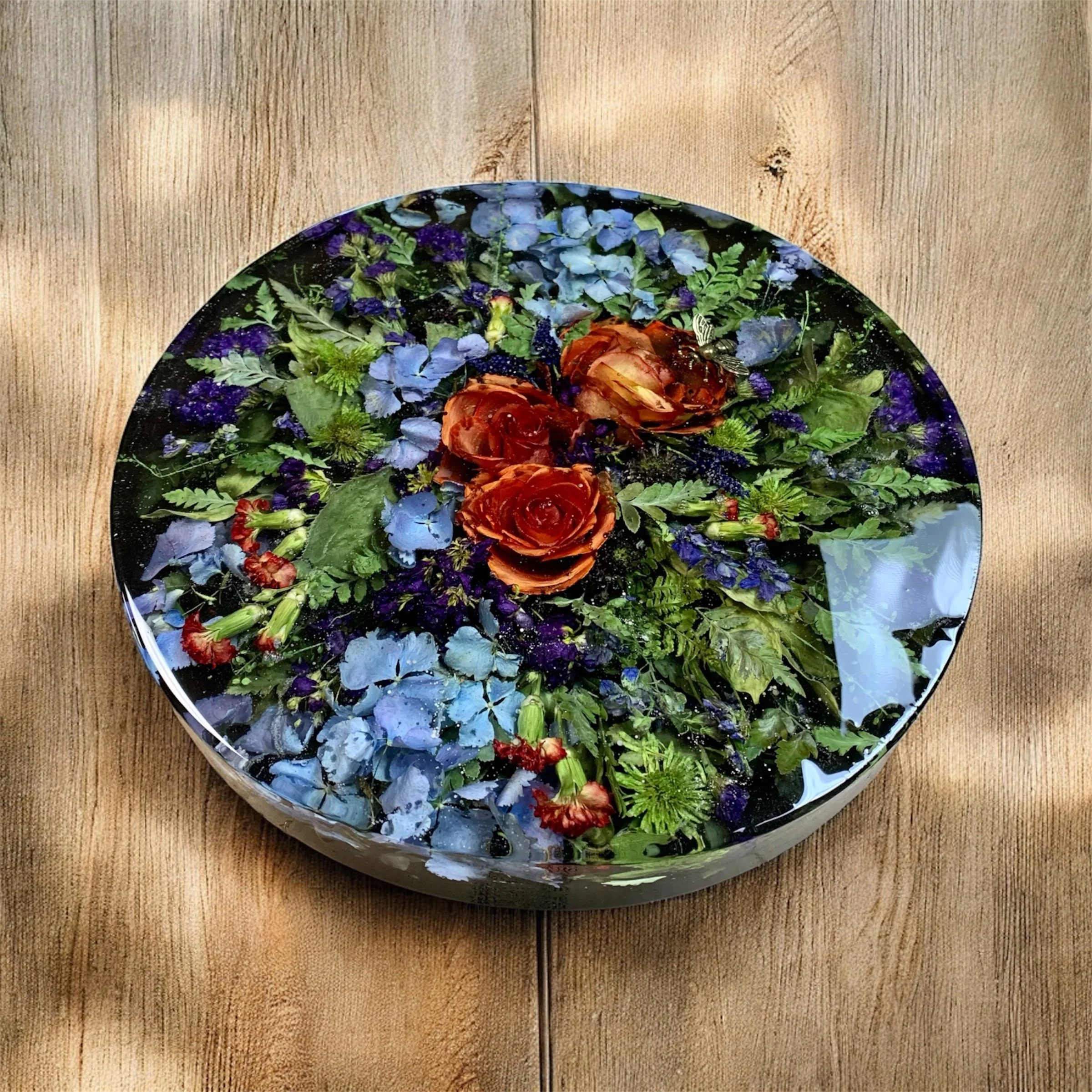 A round decorative tray filled with assortedpreserved with resin flowers and greenery, including roses and ferns.