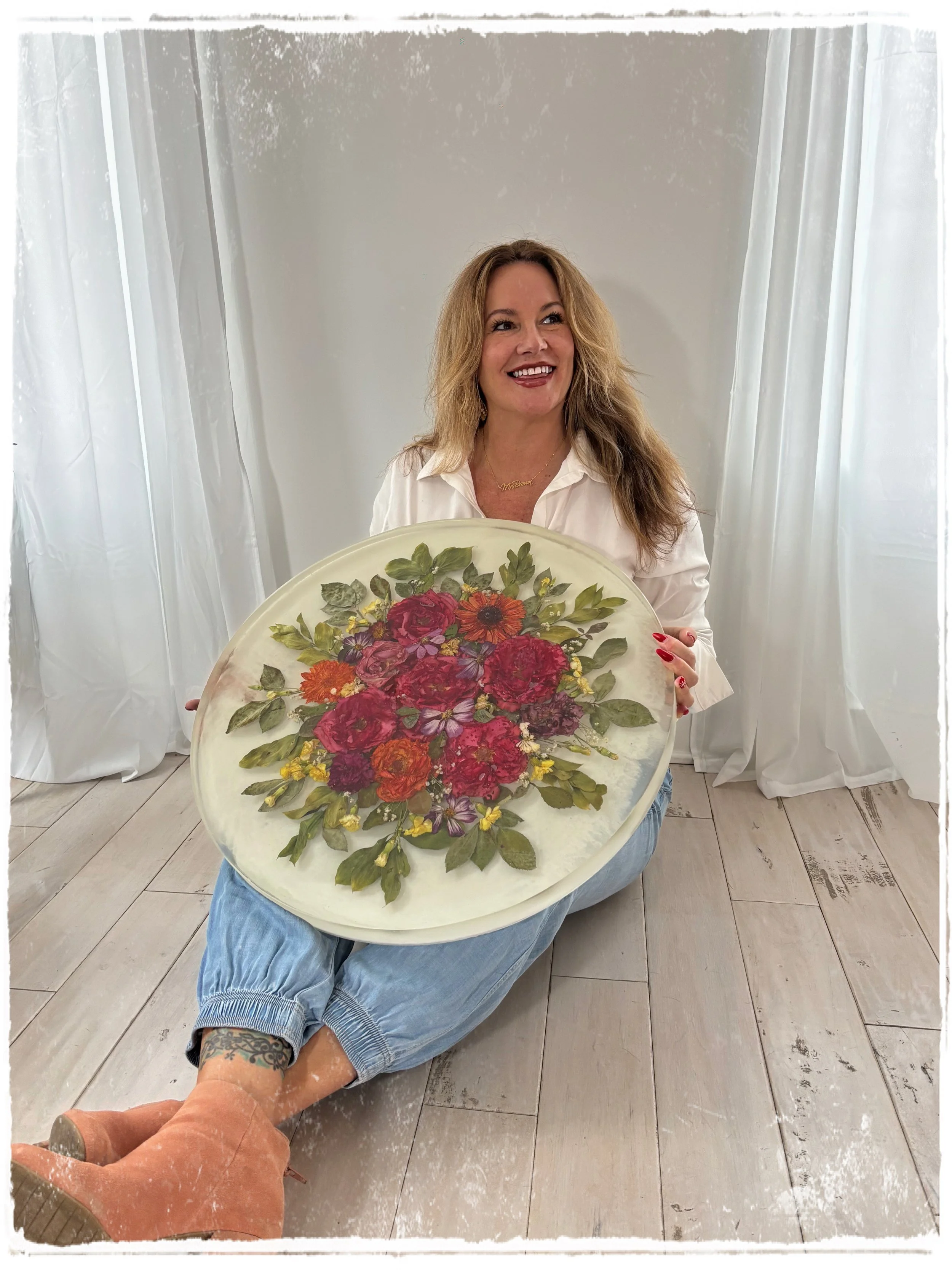 A smiling woman with blonde hair in a white shirt and jeans, sitting on the floor, holding a large tray with a floral arrangement of colorful flowers and green leaves.