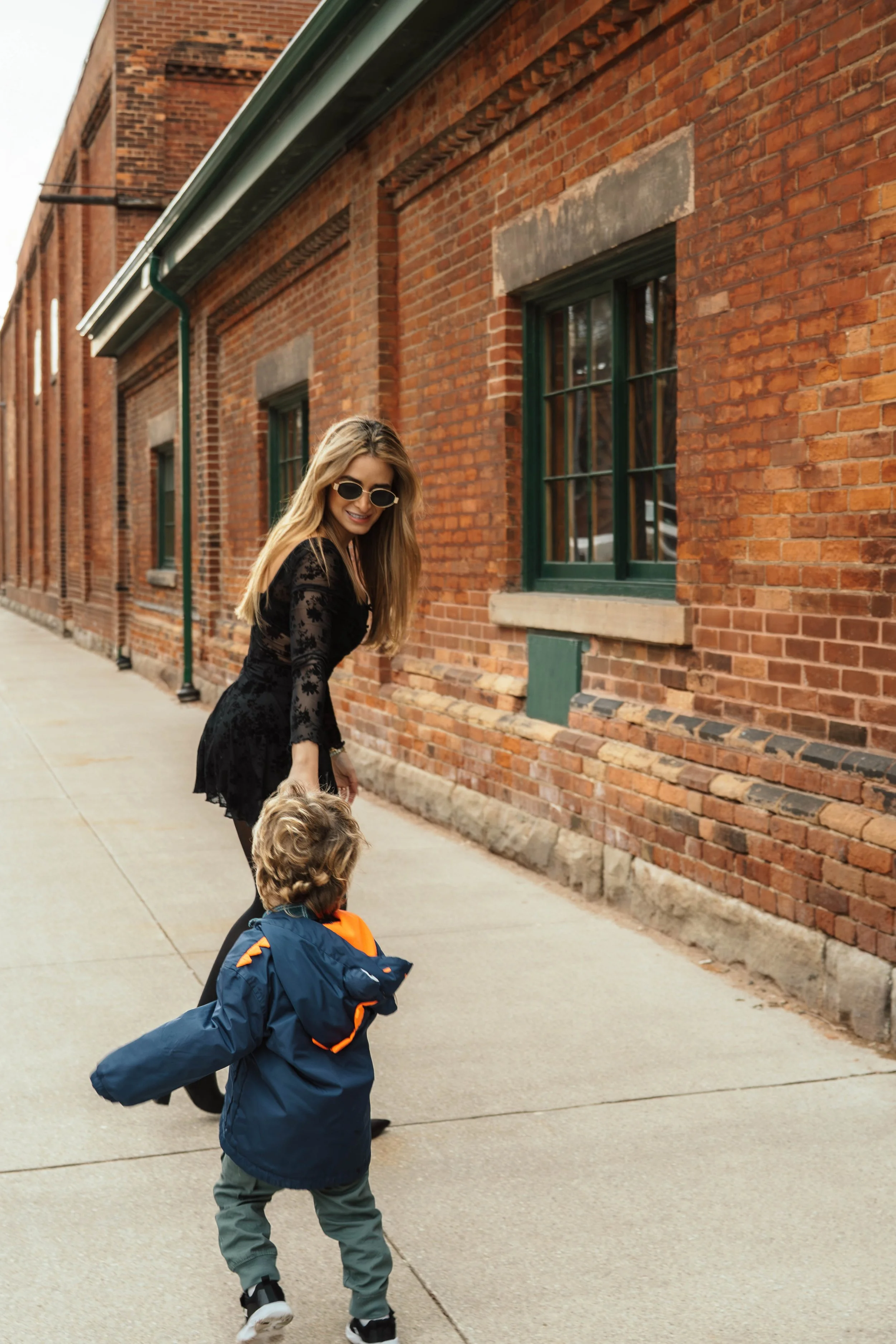 A woman with blonde hair and sunglasses smiles as she walks with two young children on a sidewalk in front of a brick building. One child wears a blue jacket and the other is partially visible, wearing a dark jacket with orange accents.