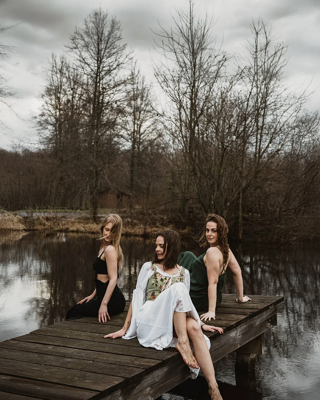 Three women sitting on a wooden dock by a lake or pond, surrounded by leafless trees under a cloudy sky in a natural outdoor setting.