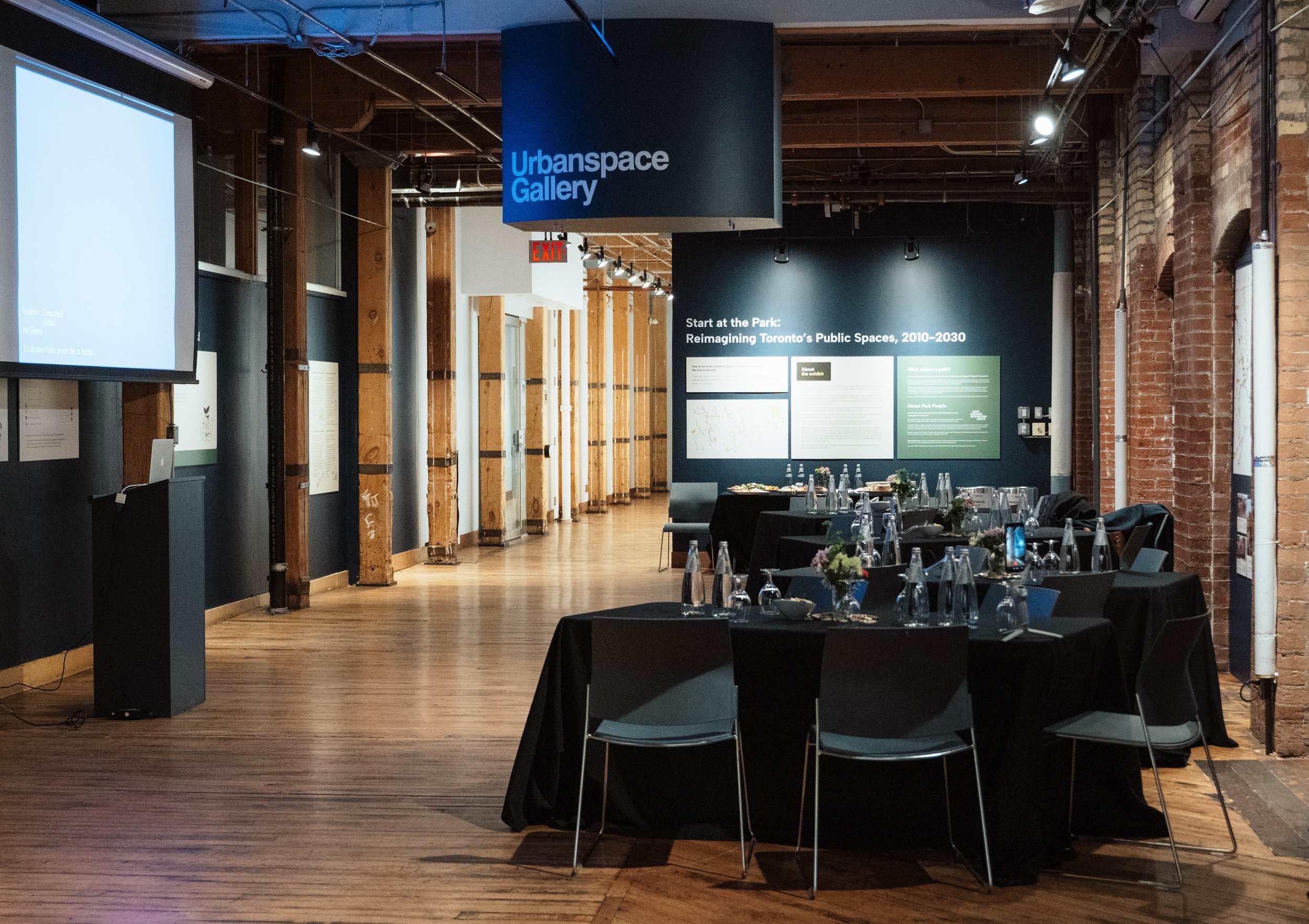 An indoor exhibition space titled 'Urbanspace Gallery' with a table set up for a meeting or event, decorated with flowers and bottles, and a large screen on the left side displaying information.