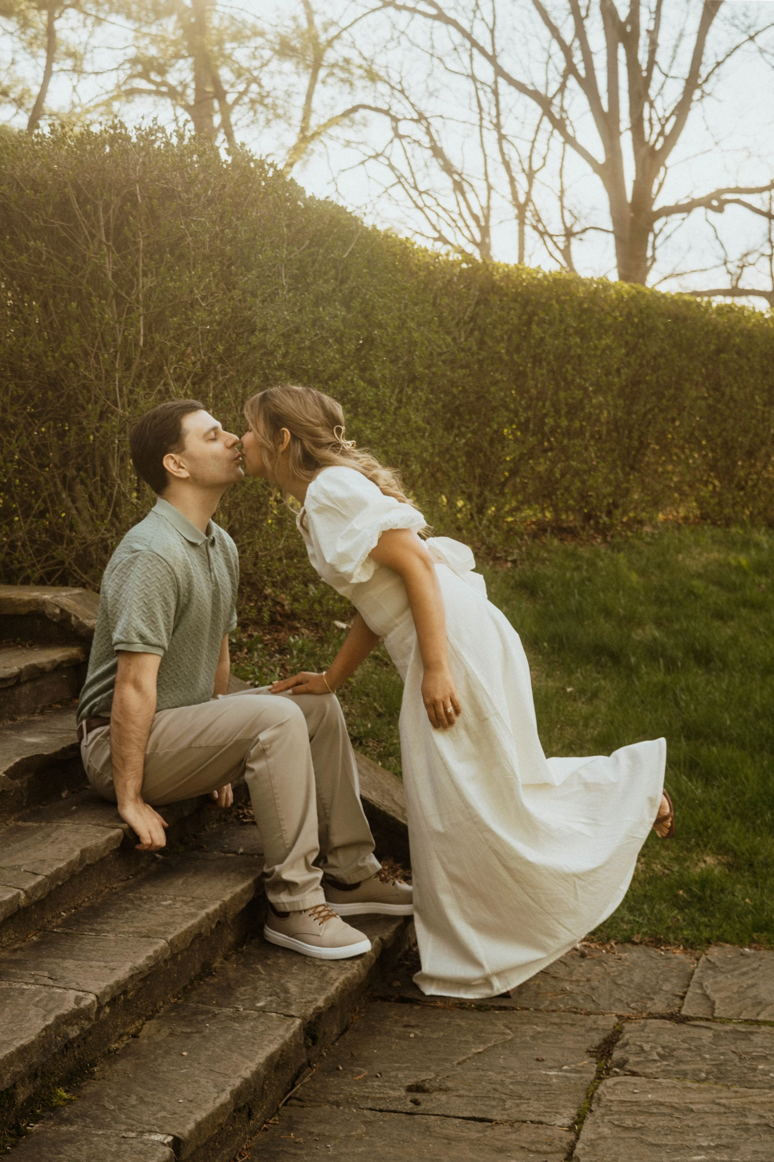 A young couple shares a kiss outdoors on stone steps, with a woman leaning over a seated man, in front of a green hedge and trees with bare branches.