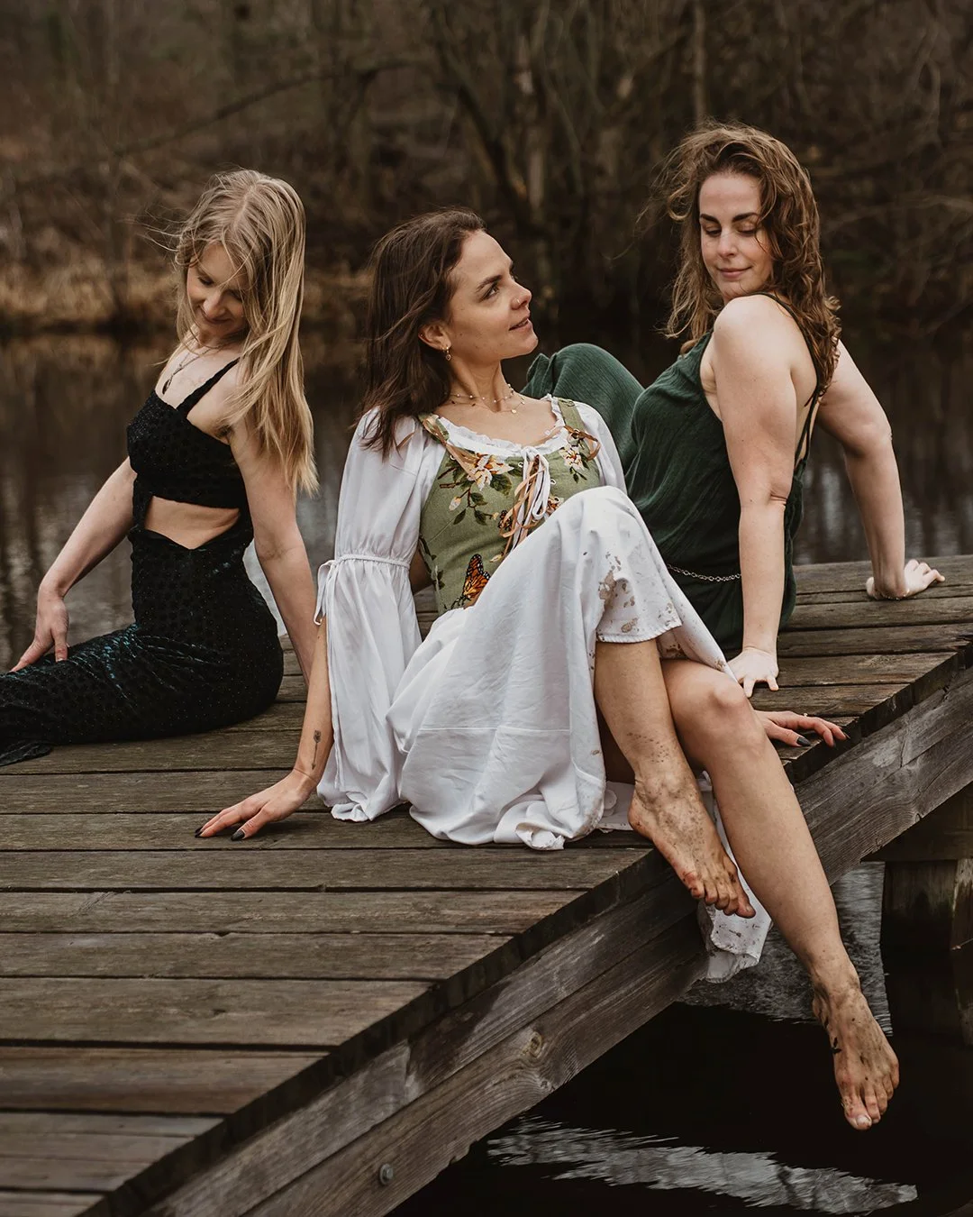 Three women sitting on a wooden dock by a body of water surrounded by trees.
