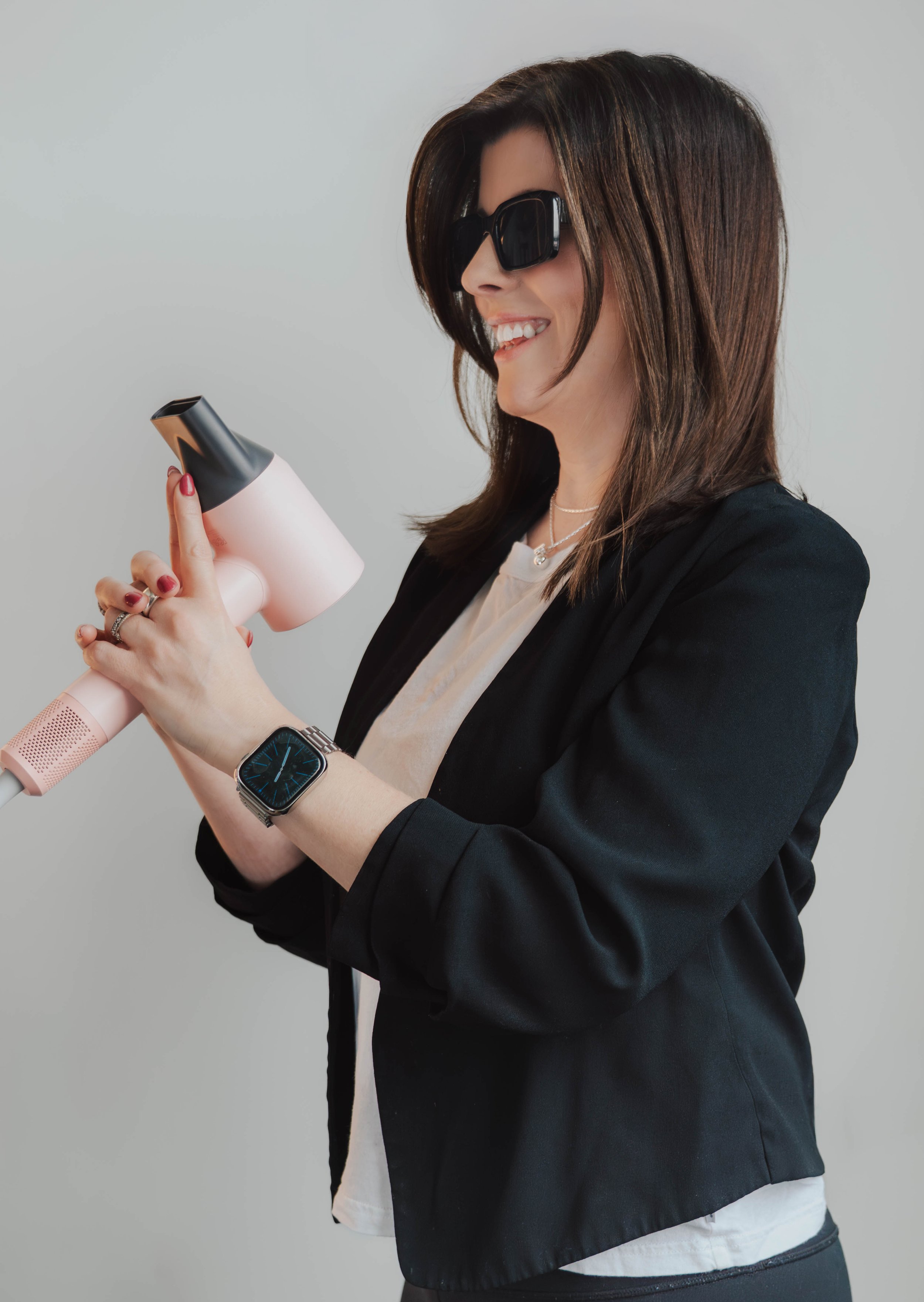 A woman with shoulder-length brown hair, wearing black sunglasses, a black blazer, and a watch, smiling while holding a pink hair dryer.