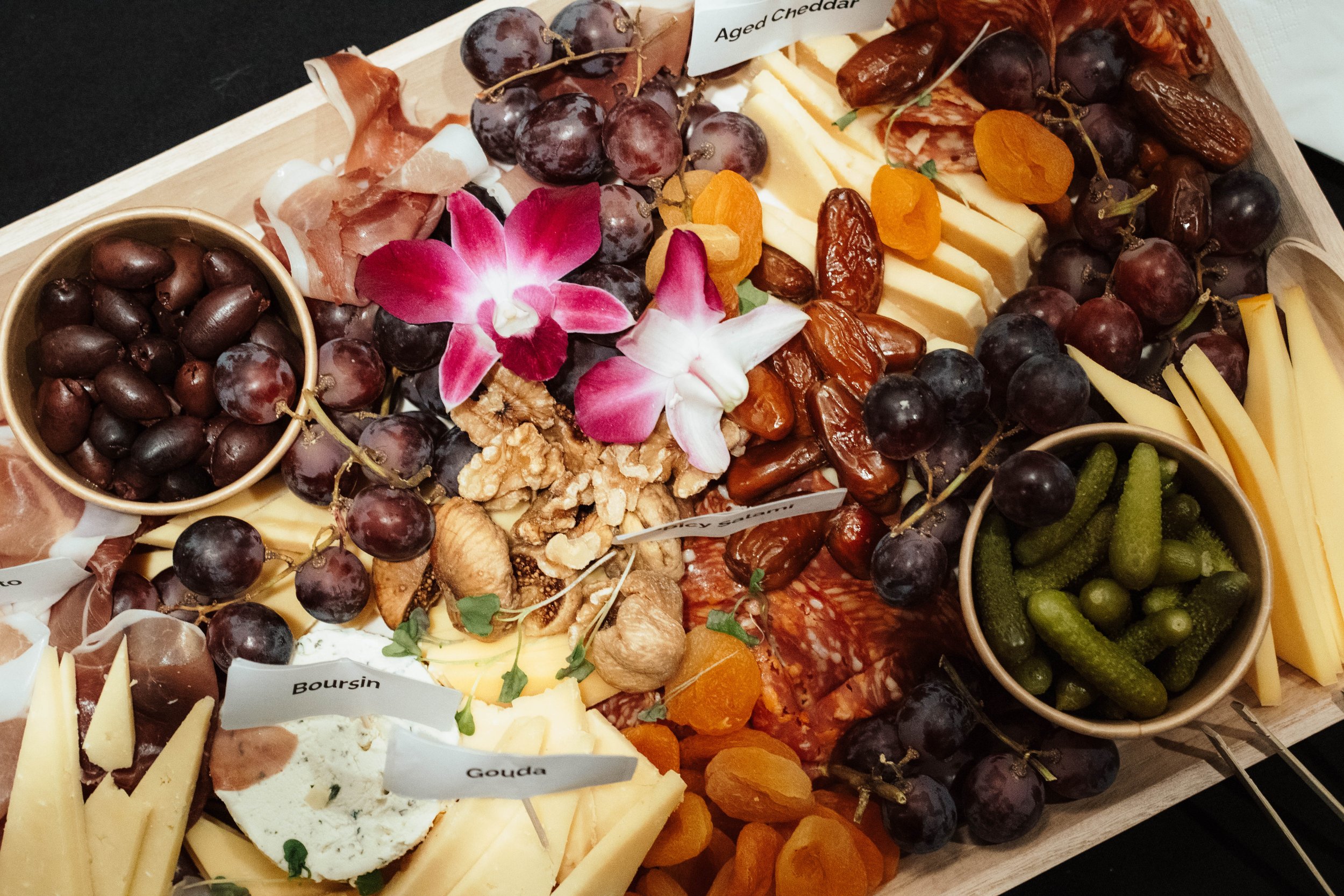 A cheese and fruit platter featuring grapes, cured meats, cheese slices labeled Gouda and Boursin, dried apricots, pickles, and decorative flowers.