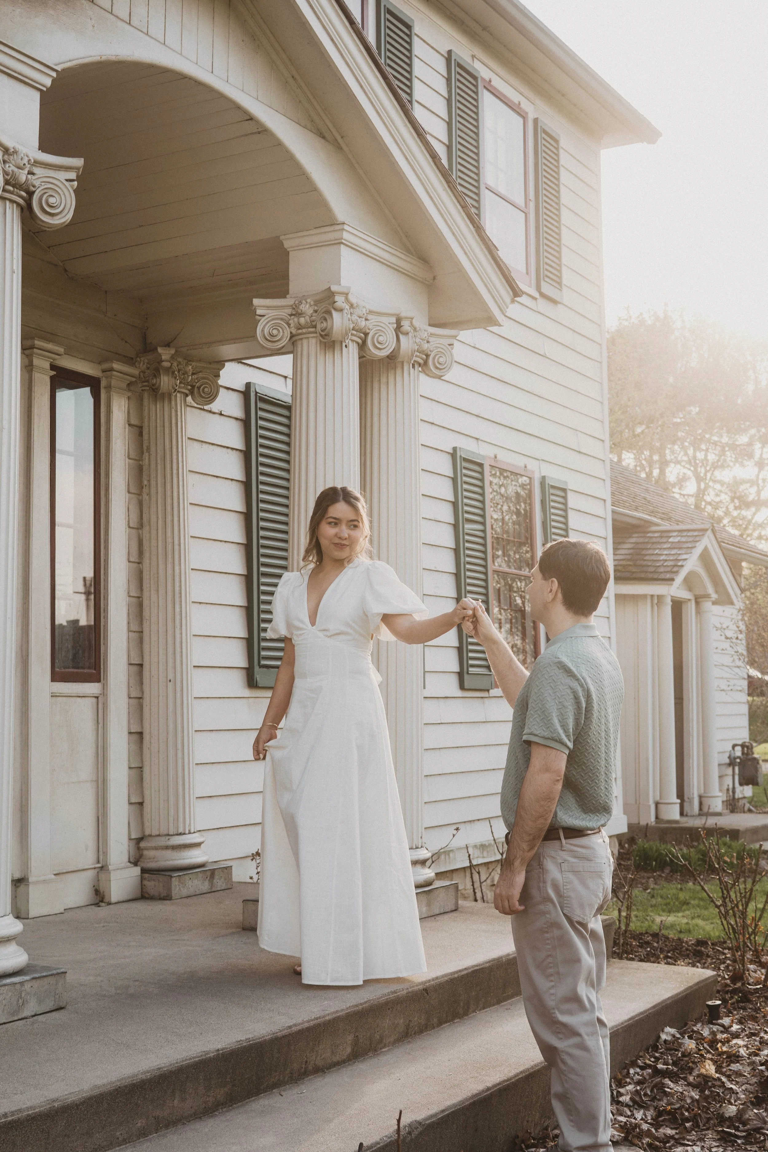 A woman in a white dress is being proposed to by a man in a gray shirt and beige pants on the porch of a white, classic-style house with columns and green shutters.