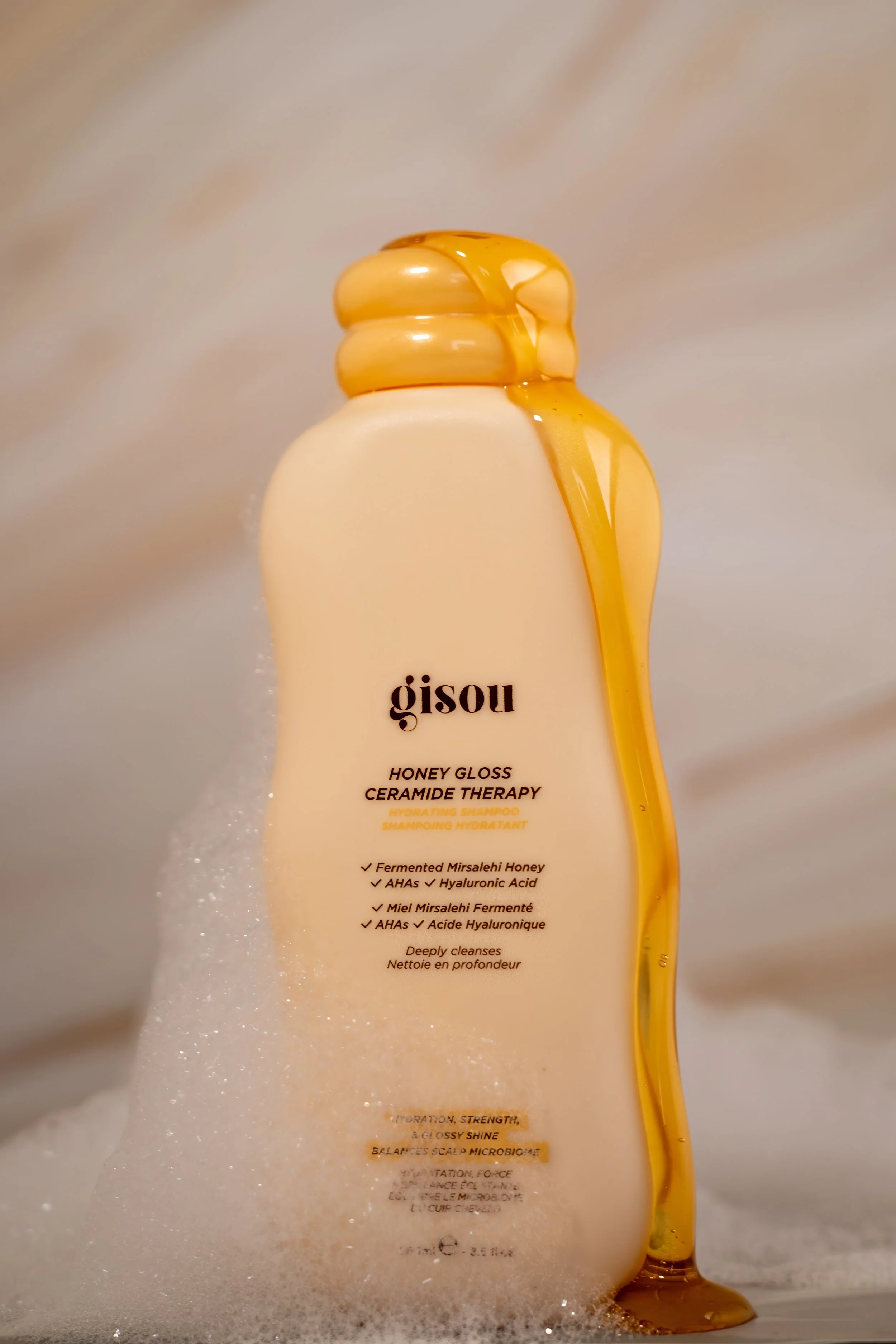 A bottle of Gisou Honey Gloss Ceramide Therapy Hydrating Shampoo with honey dripping down the side, surrounded by foam and bubbles.