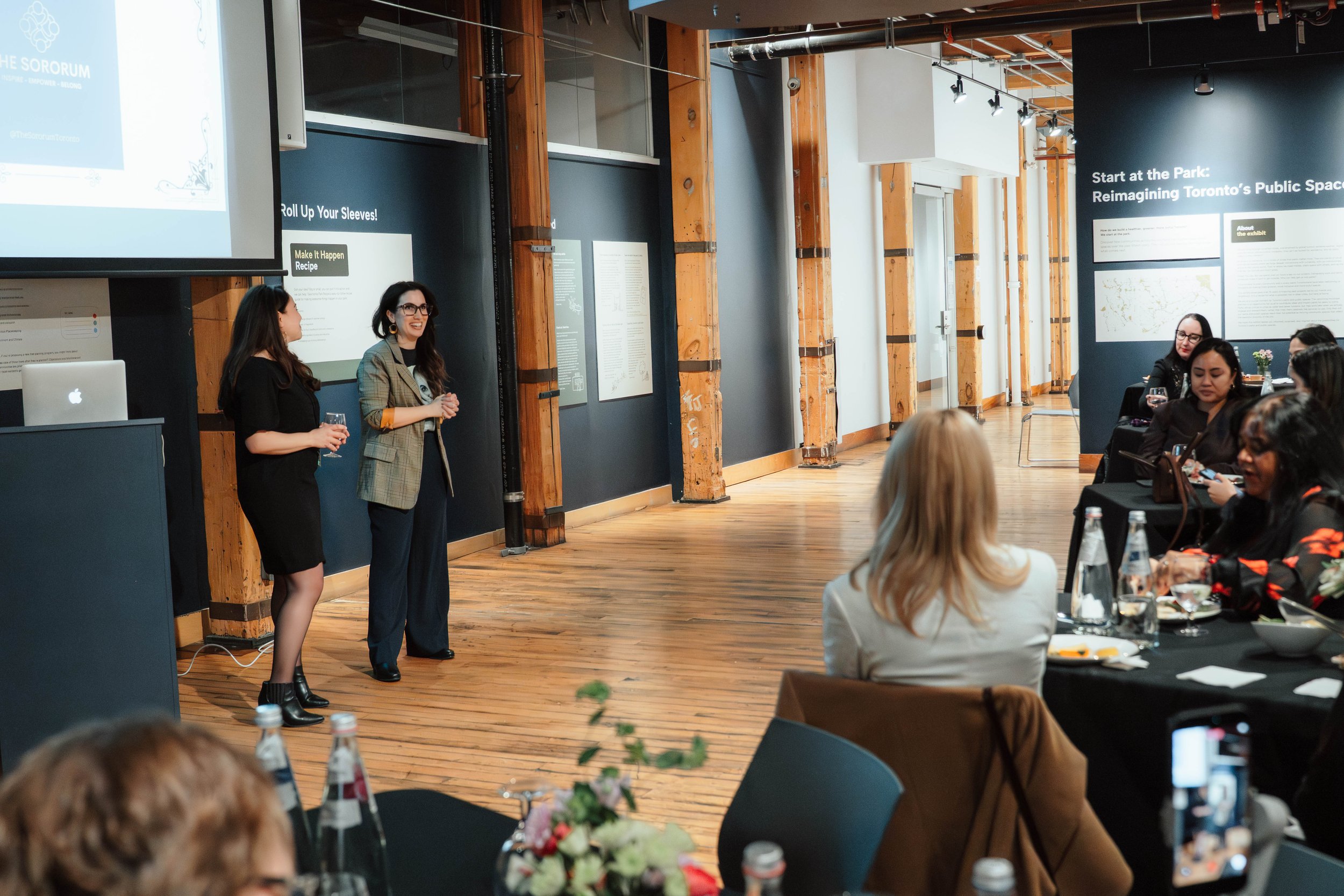 Women giving a presentation in front of an audience in a modern conference room with wooden floors and dark walls.