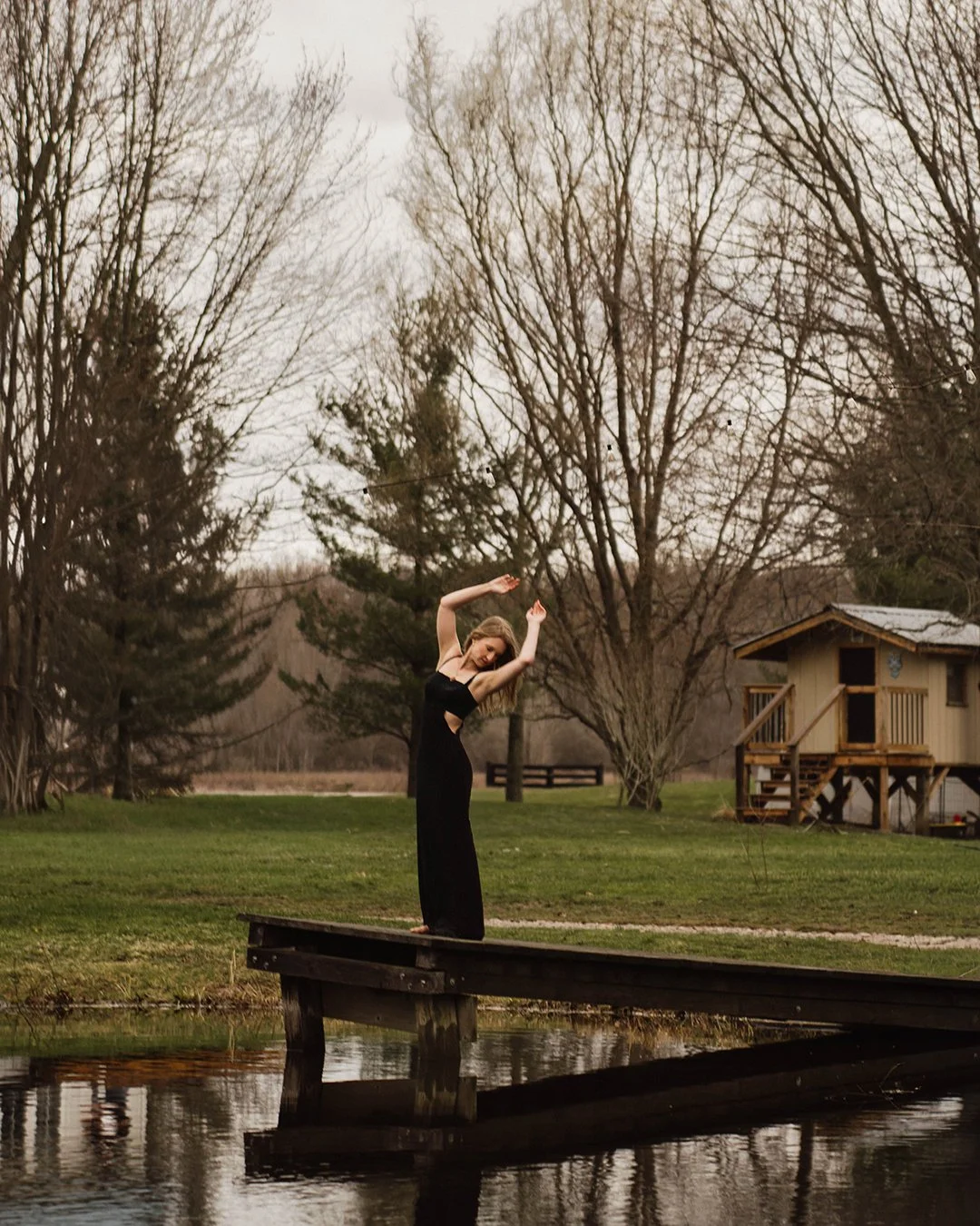 A woman in a black dress standing on a wooden dock by a pond, surrounded by trees with no leaves, during overcast weather.