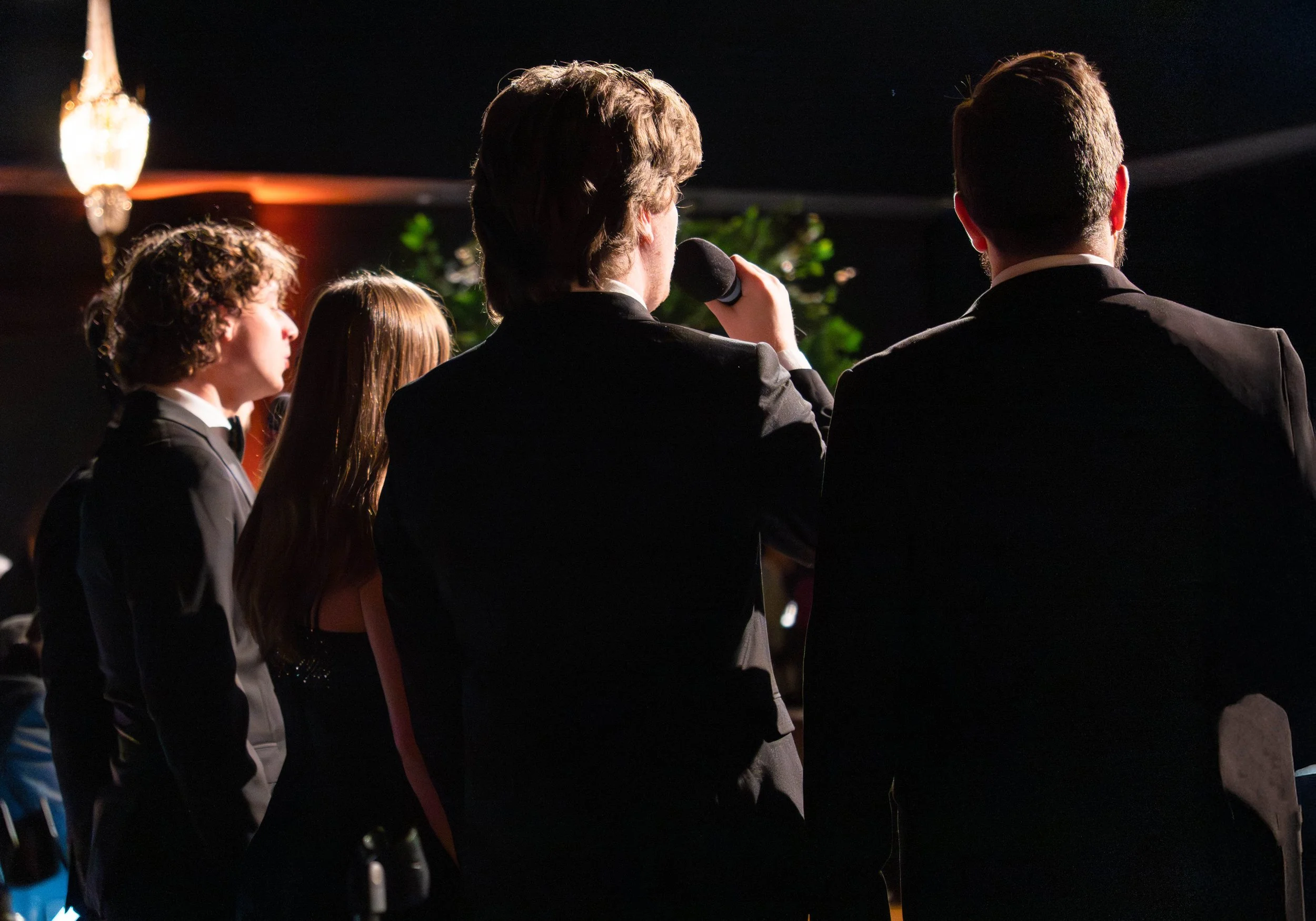 Group of people in formal attire, with one person holding a microphone, seen from behind, in a dimly lit setting with a chandelier and some greenery in the background.