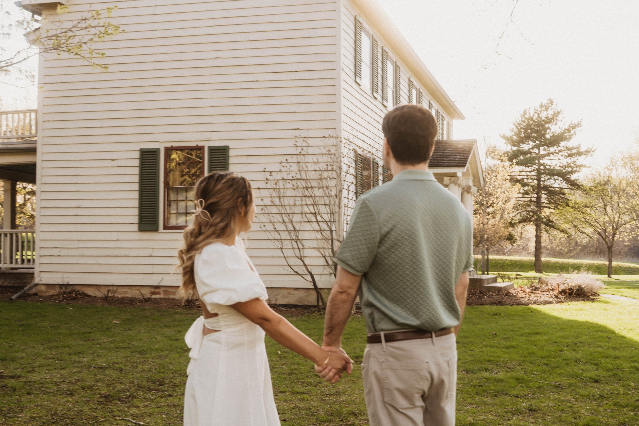A young man and woman holding hands and standing outdoors in front of a large house, with the woman looking at the man. The house is a white wooden structure with multiple windows and green shutters, surrounded by trees and a well-kept yard.