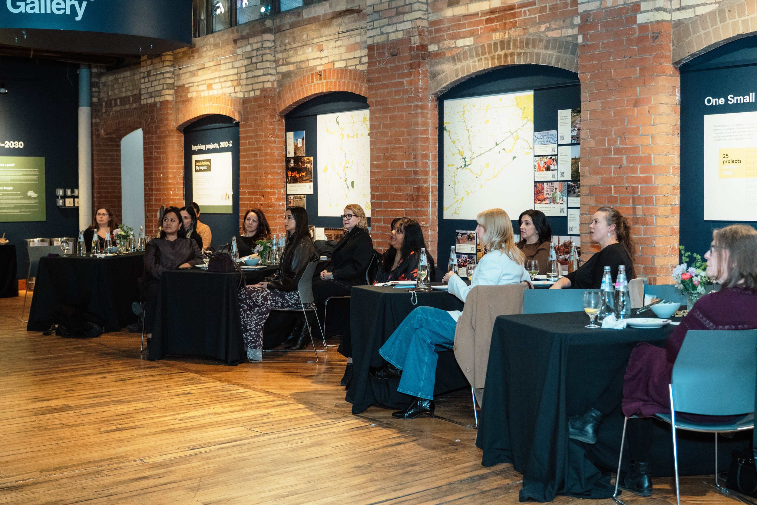 A group of women seated at a long banquet-style table, attending a presentation in a brick-walled gallery space, with informational posters and maps displayed on the walls behind them.