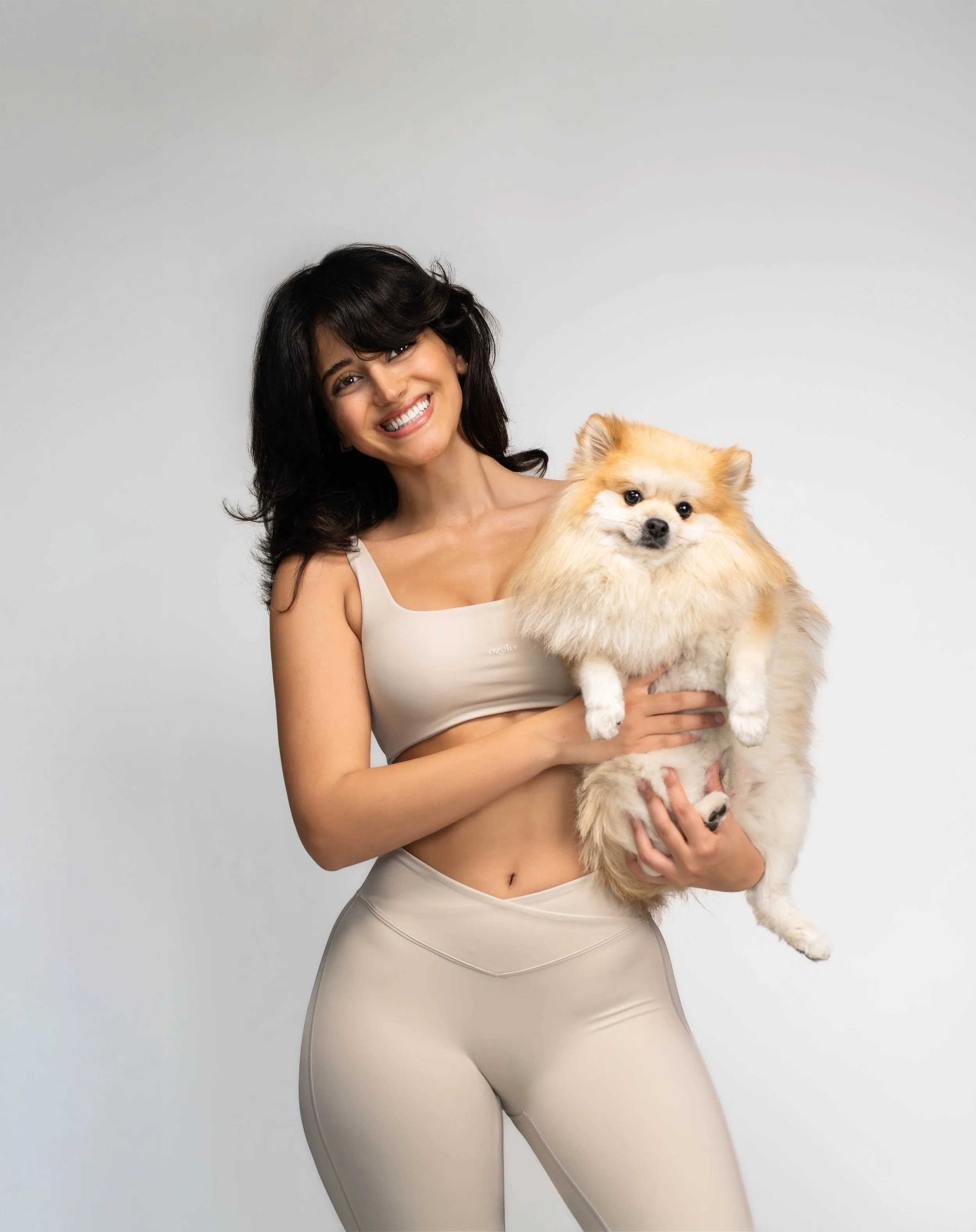 A woman in a beige sports bra and matching leggings holding a fluffy Pomeranian dog, both smiling at the camera against a plain gray background.