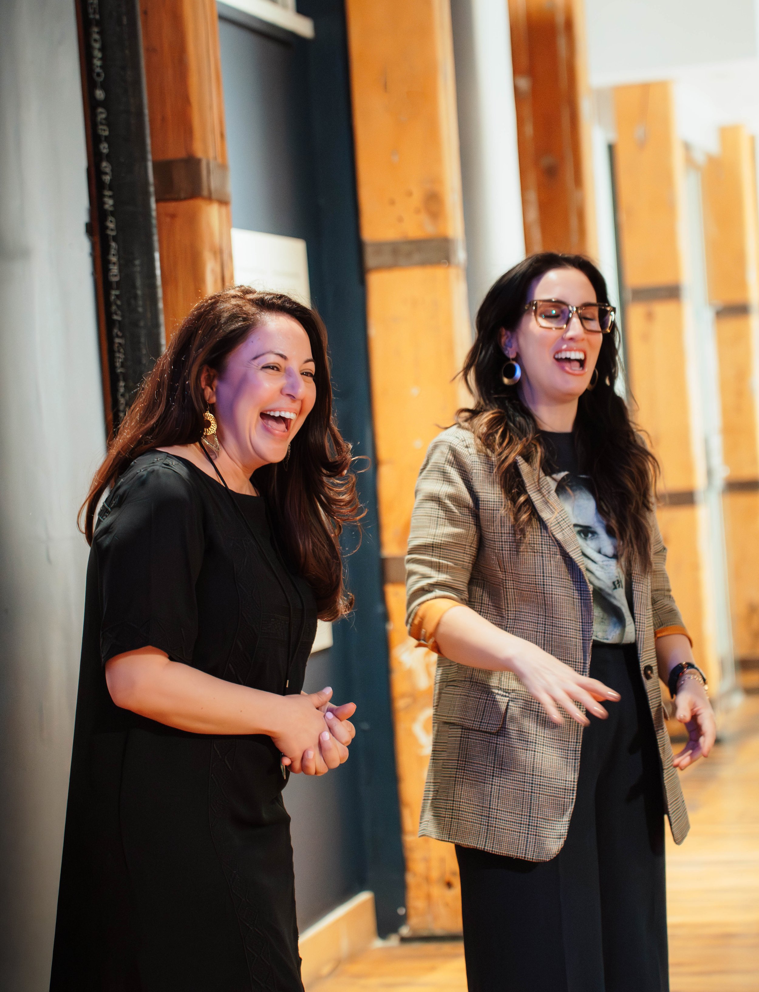 Two women are standing and laughing in an indoor setting with wooden paneling and a dark-colored wall.