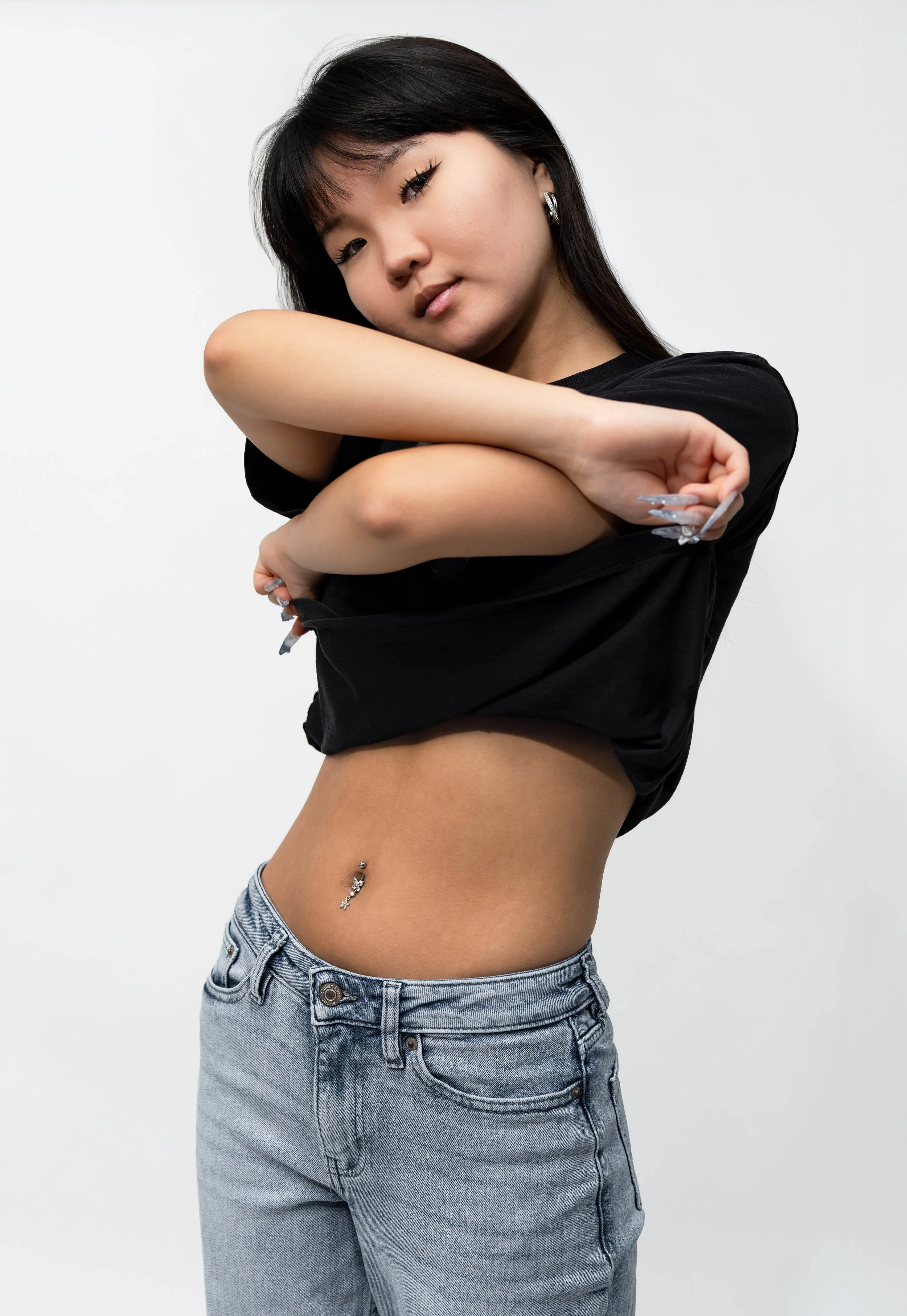 Young woman with black hair and light makeup, wearing hoop earrings, a black shirt, and light blue jeans, standing against a plain white background with her arms crossed and a slight smile.