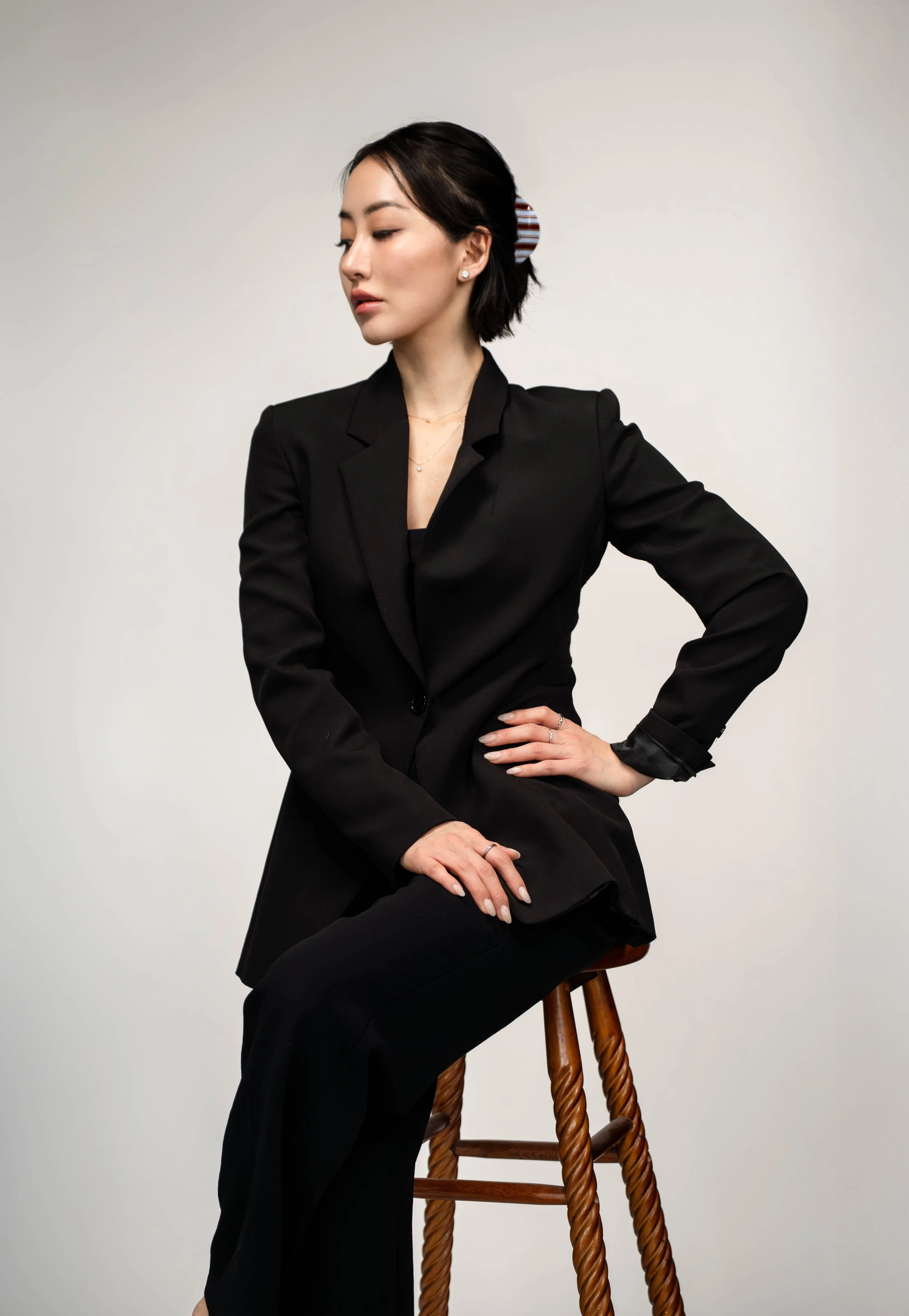 A woman sitting on a wooden stool with her eyes closed, wearing a black blazer and black pants, with short dark hair and a striped hair accessory.