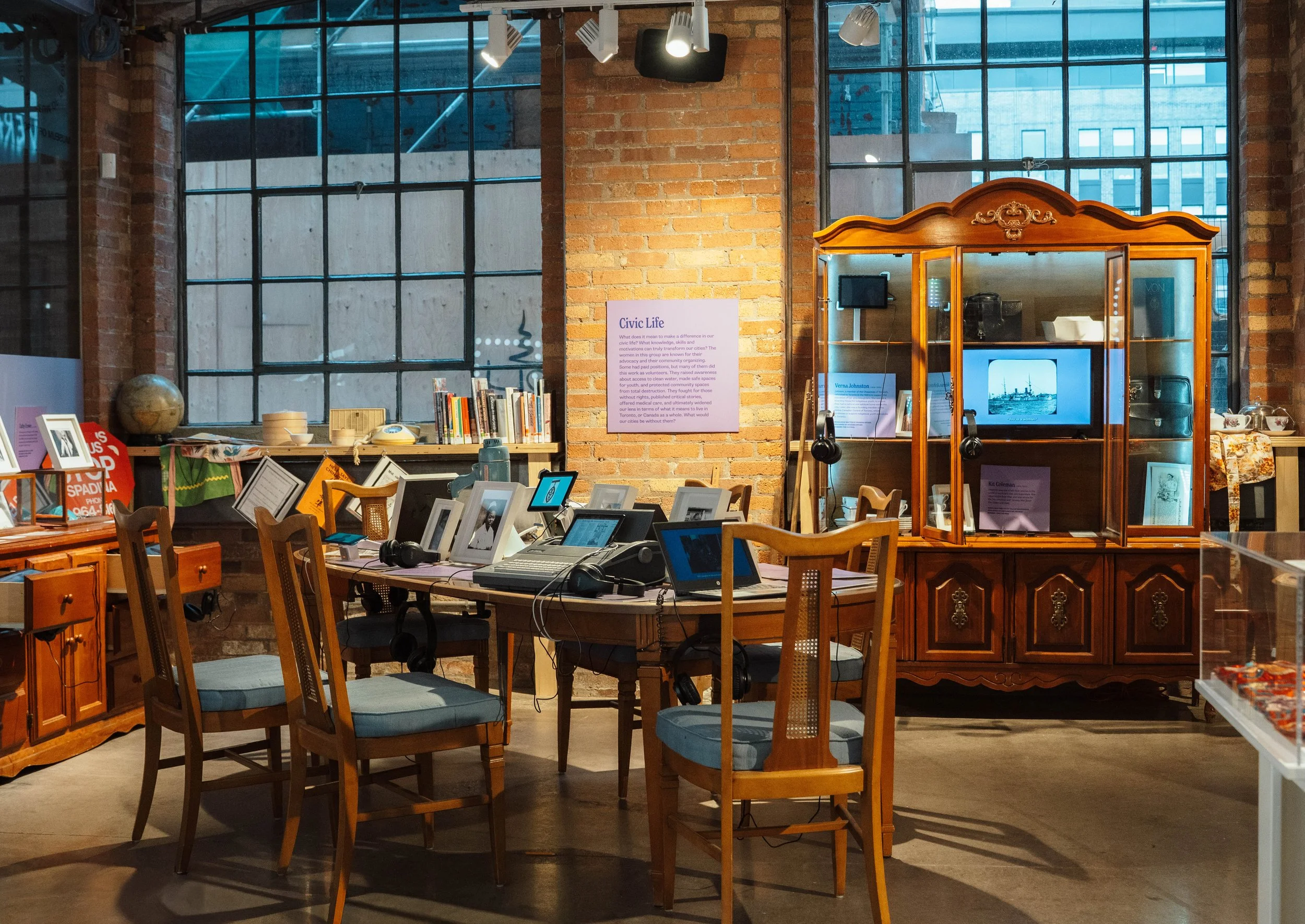 An interior view of a cozy room with brick walls and large industrial windows. The room features a wooden dining table with multiple laptops and headphones, surrounded by chairs. A vintage wooden display cabinet holds monitors and various items, with