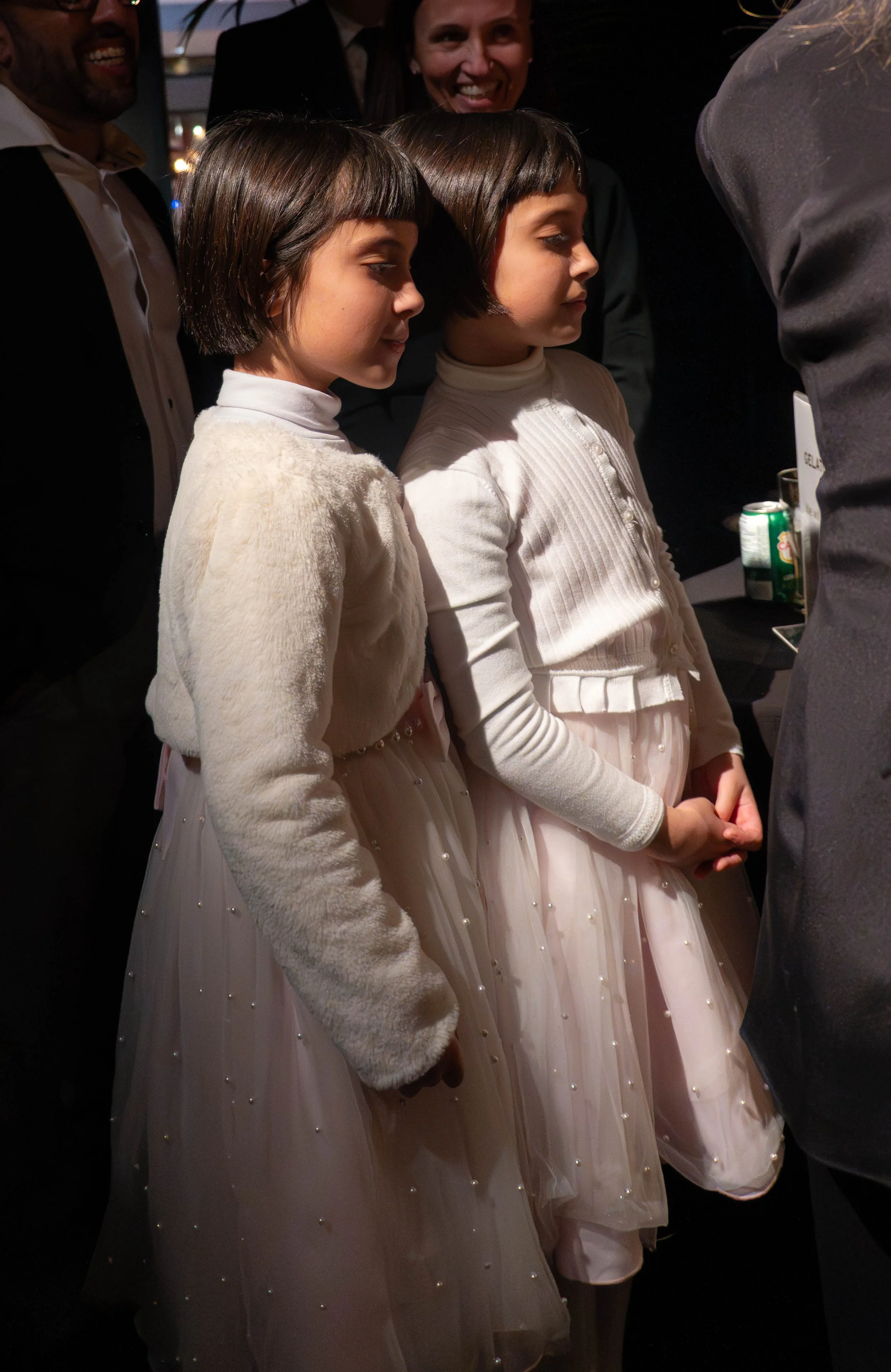 Two young girls with bob hairstyles wearing white dresses and cardigans, standing side by side, with adults around them during an indoor event.