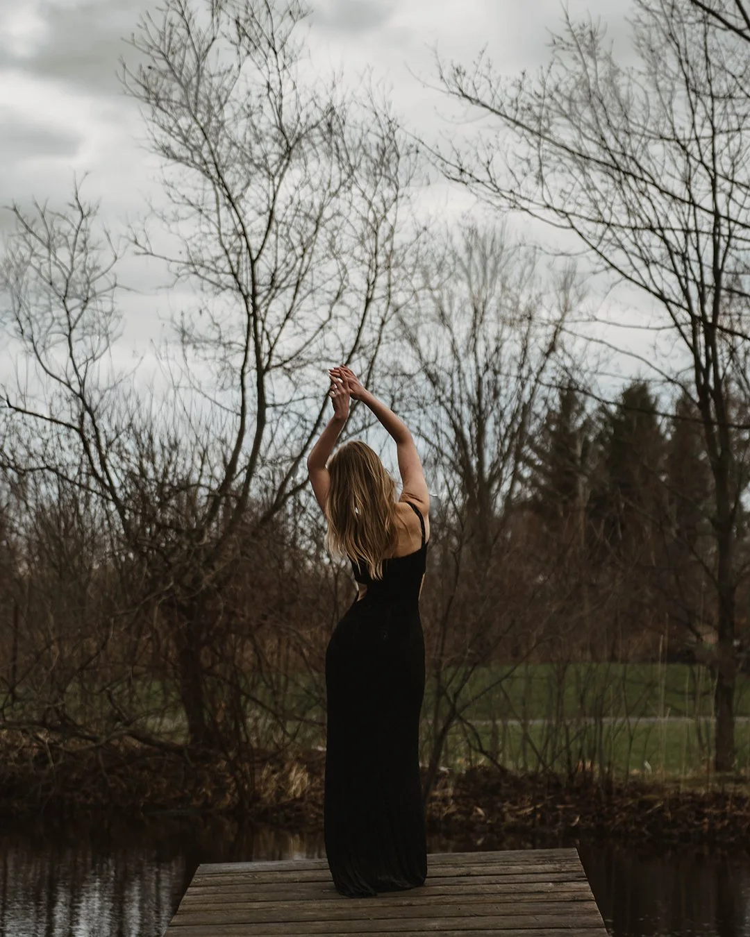 A woman in a long black dress standing on a wooden dock by a body of water, with her arms raised and hands clasped above her head. She is facing away from the camera, with leafless trees and a cloudy sky in the background.