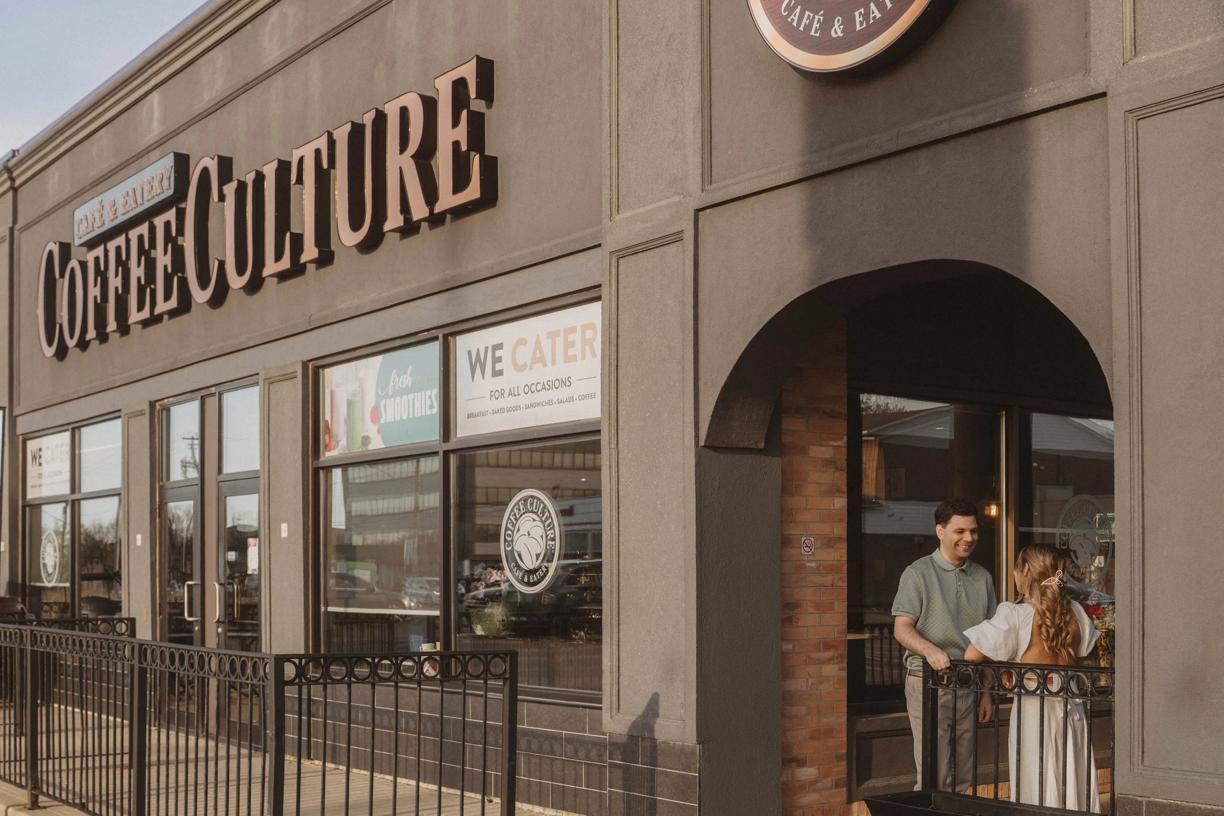 Exterior of a coffee shop with large signage reading "Coffee Culture" and smaller signs indicating catering and smoothies, two people standing at the entrance smiling and talking.