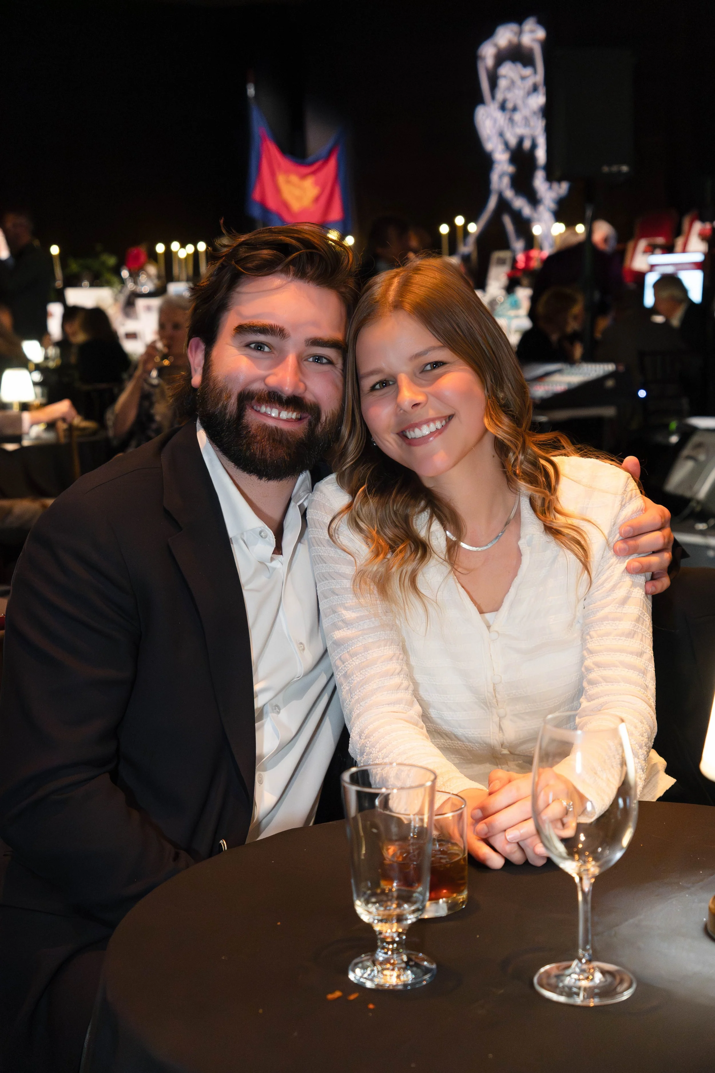 A man and a woman sitting at a table, smiling and embracing at a formal event or celebration, with a blurred background featuring other guests and decorations.