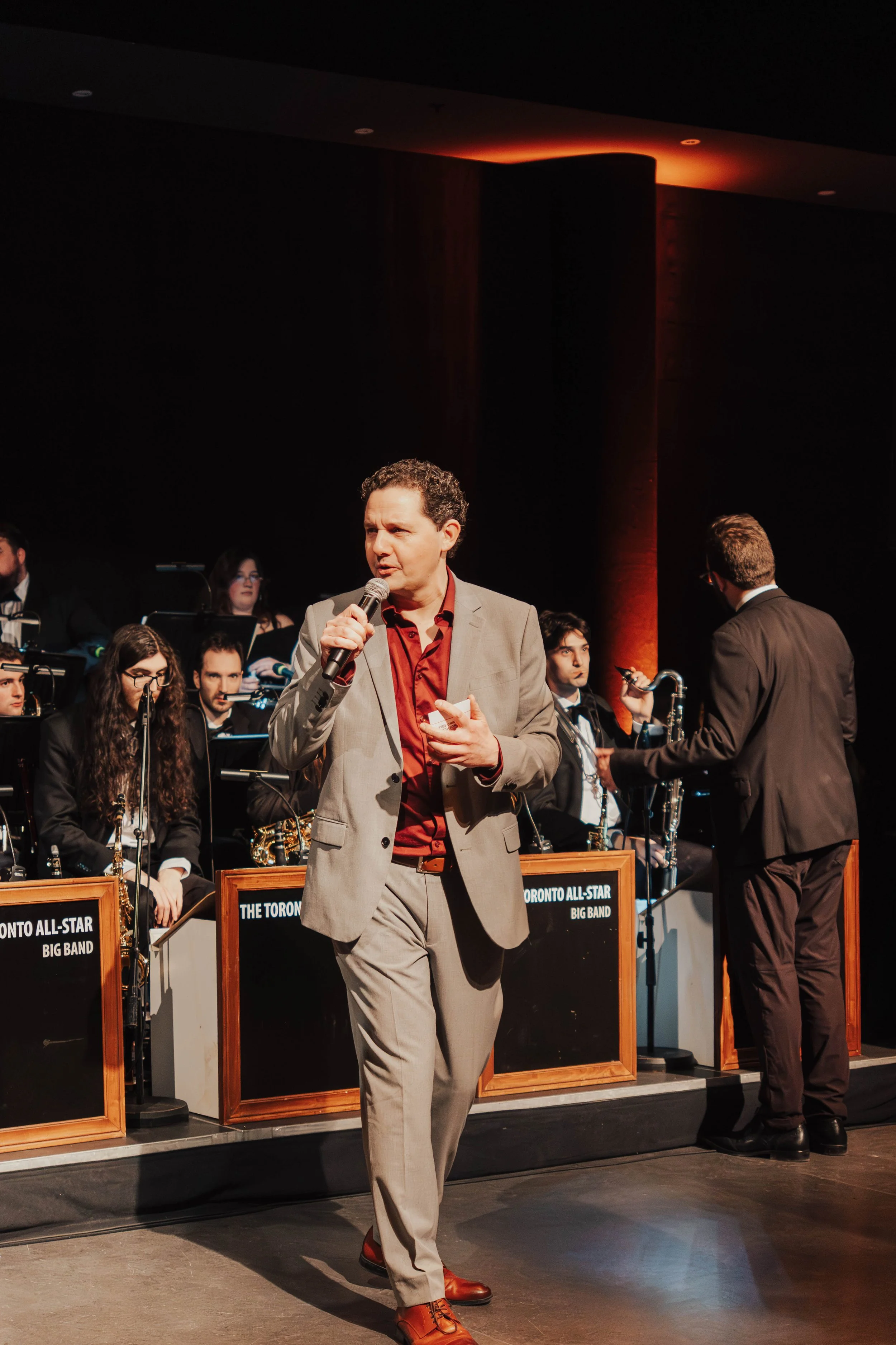 A man in a light gray suit and red shirt holding a microphone and speaking at a formal event with a band in the background.
