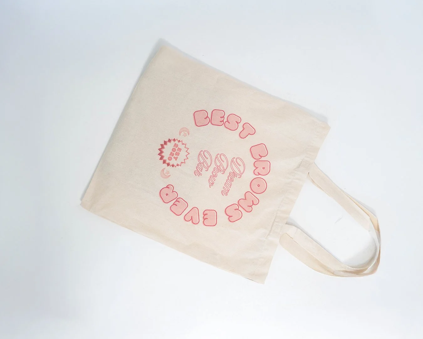Cream-colored tote bag with pink text and design promoting a clothing brand, laying flat on a white surface.