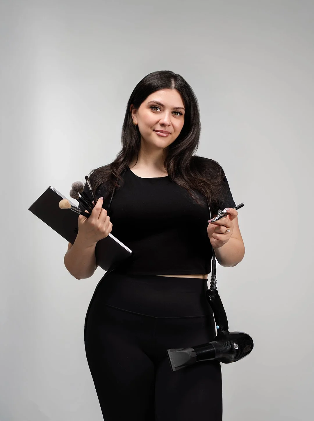 A woman holding makeup brushes in her left hand and a small device in her right hand, with a hairdryer hanging from her waist, standing against a plain gray background.