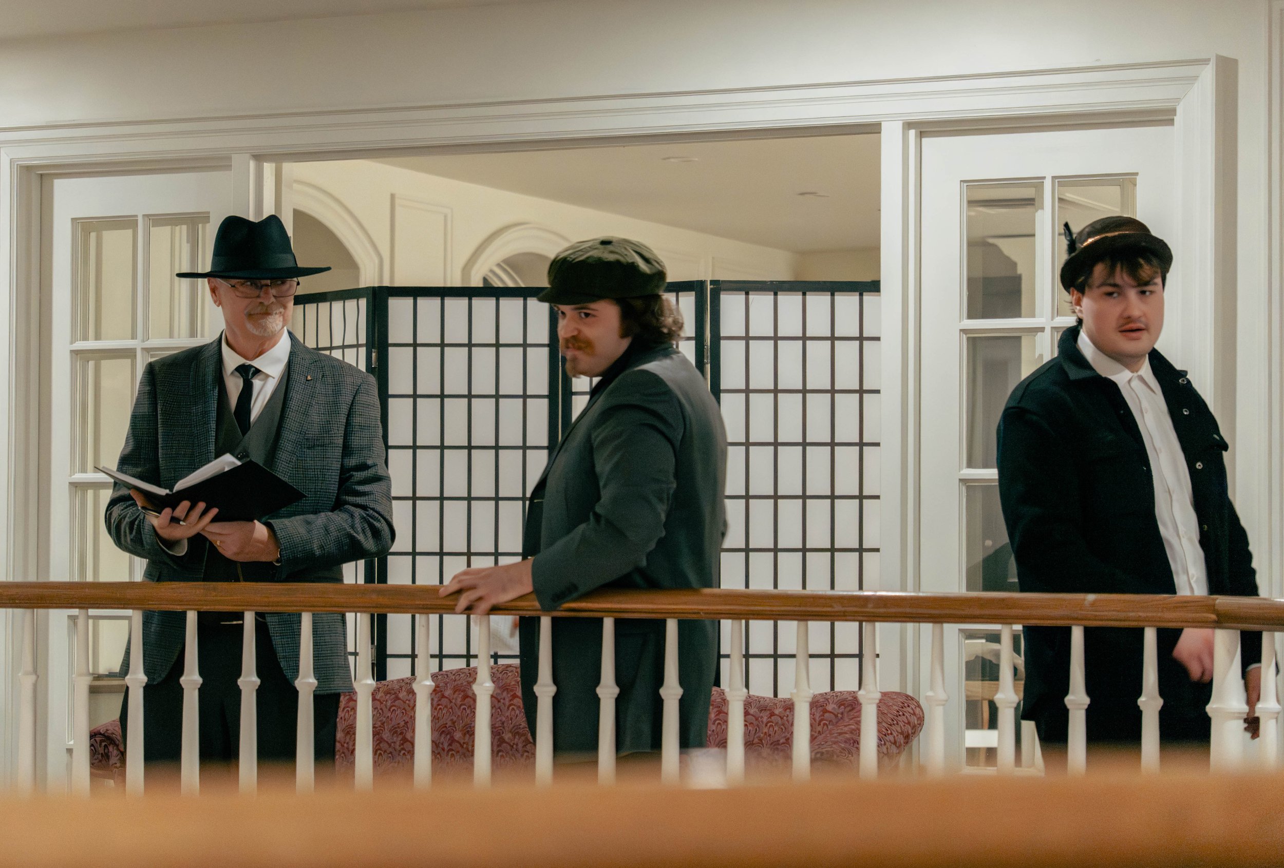 Three men standing behind a wooden railing in an elegant, well-lit room with white walls and decorative paneling. The man on the left is wearing a black hat, glasses, a checkered blazer, and a black tie. The man in the middle has a mustache, wearing 