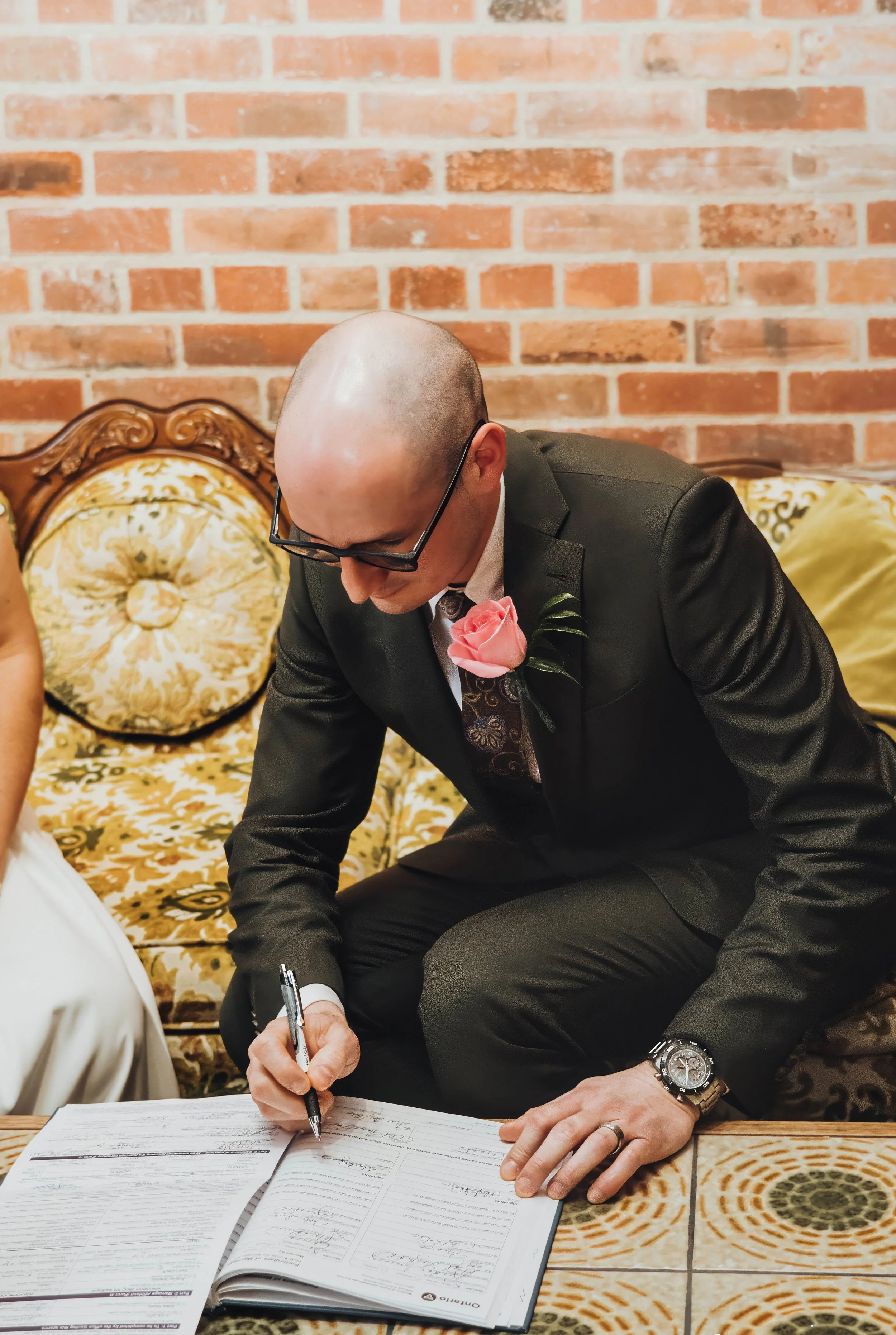 A man in a black suit with a pink rose boutonniere is signing a marriage register, sitting on a vintage floral sofa with a brick wall background.
