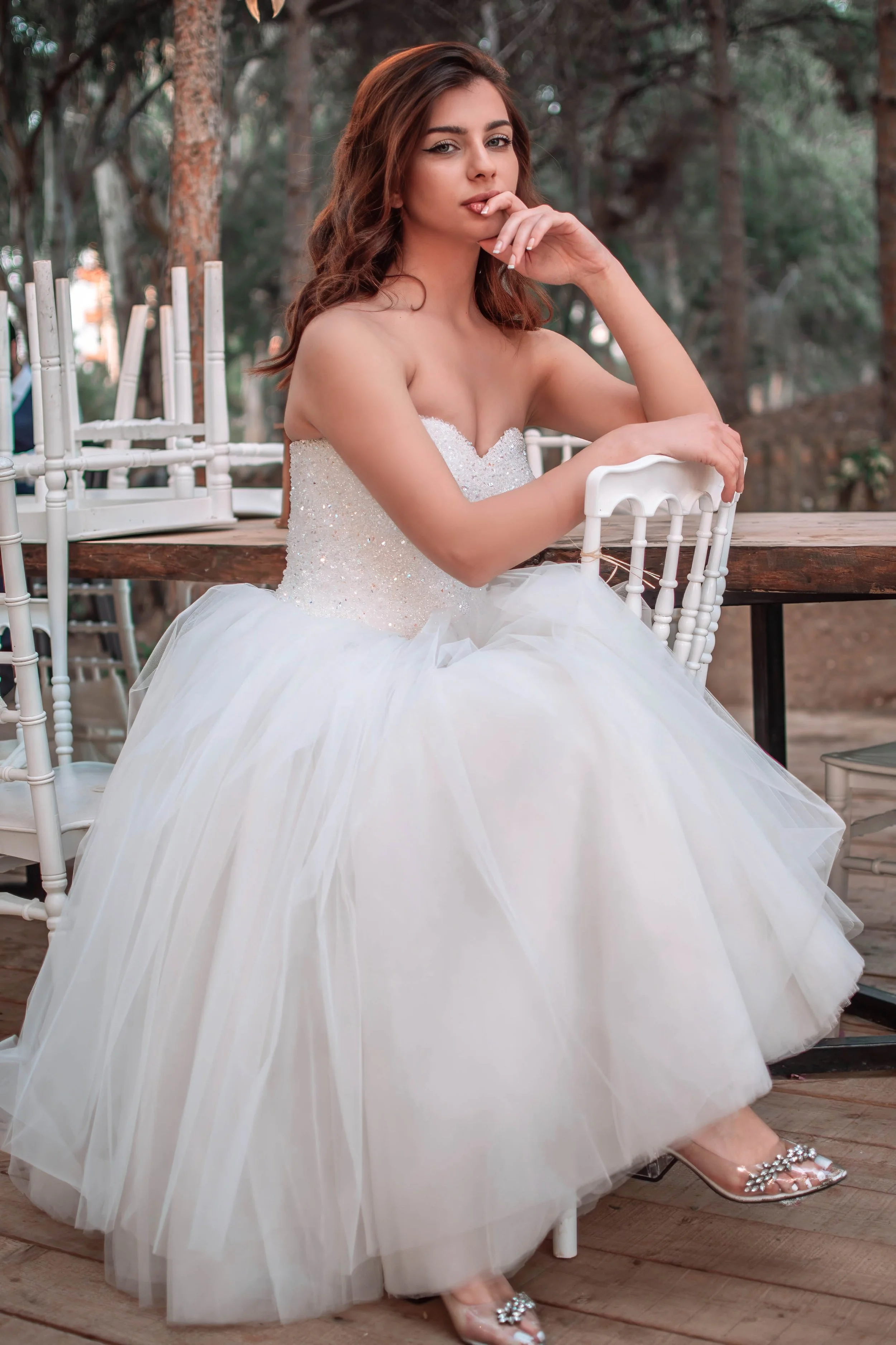 A young woman in a wedding dress with a beaded bodice and full tulle skirt sitting outdoors at a wooden table in a wooded area, with chairs stacked behind her.