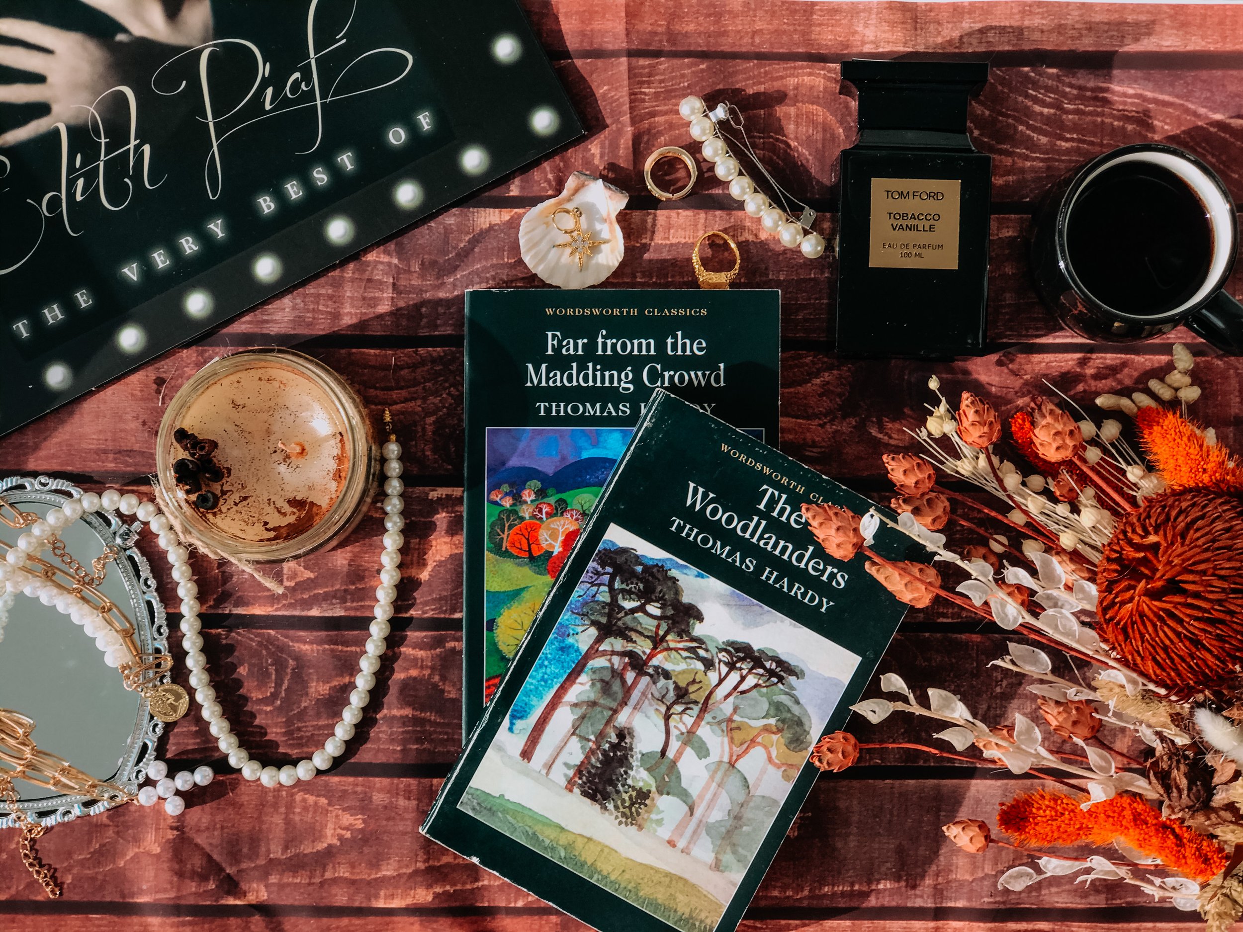 An assortment of books, jewelry, a candle, a cup of coffee, a perfume bottle, and dried flowers on a wooden surface.