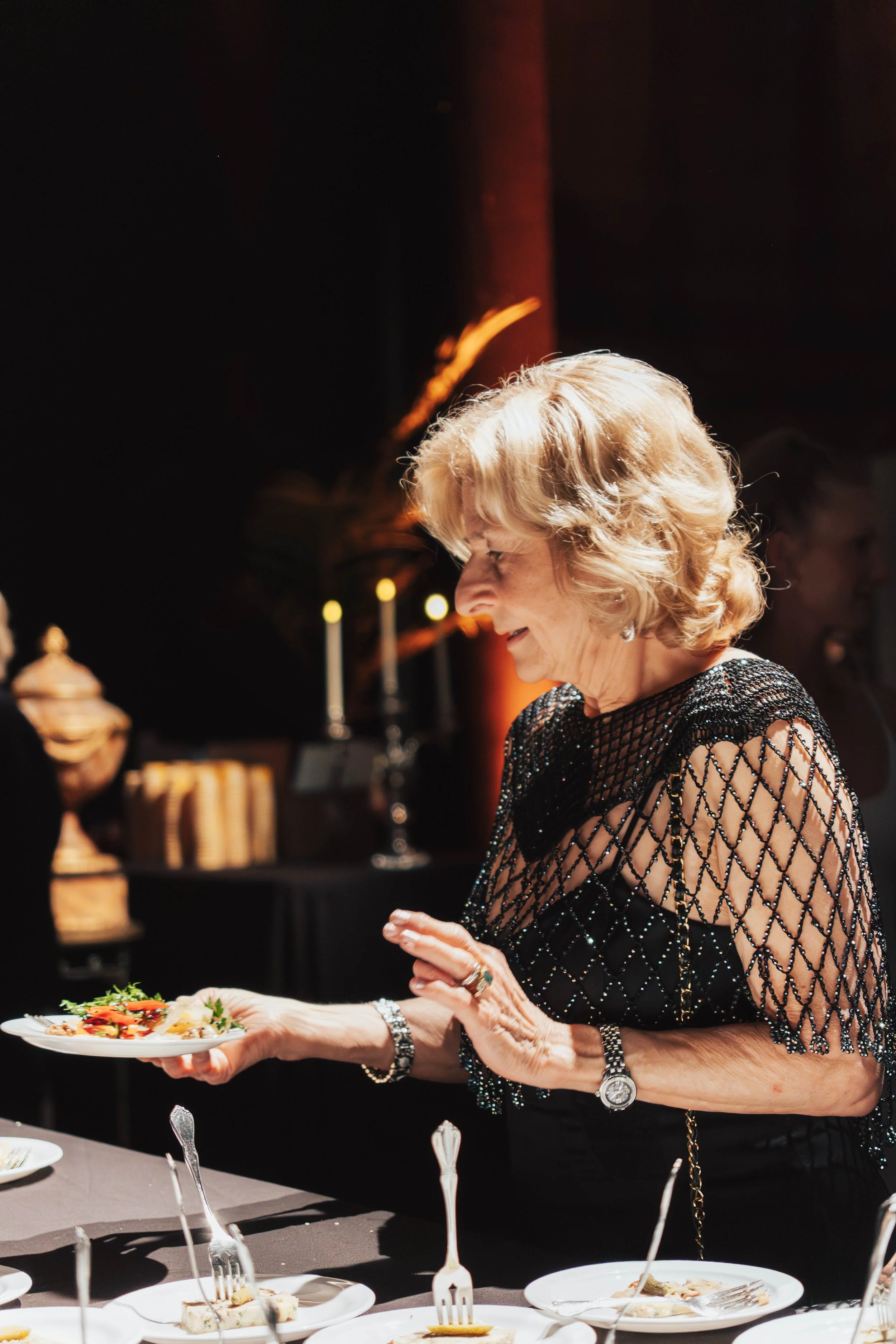 An elderly woman with blonde hair smiling and holding a plate of food at a banquet or dinner event, dressed in a black, sparkly, netted top.