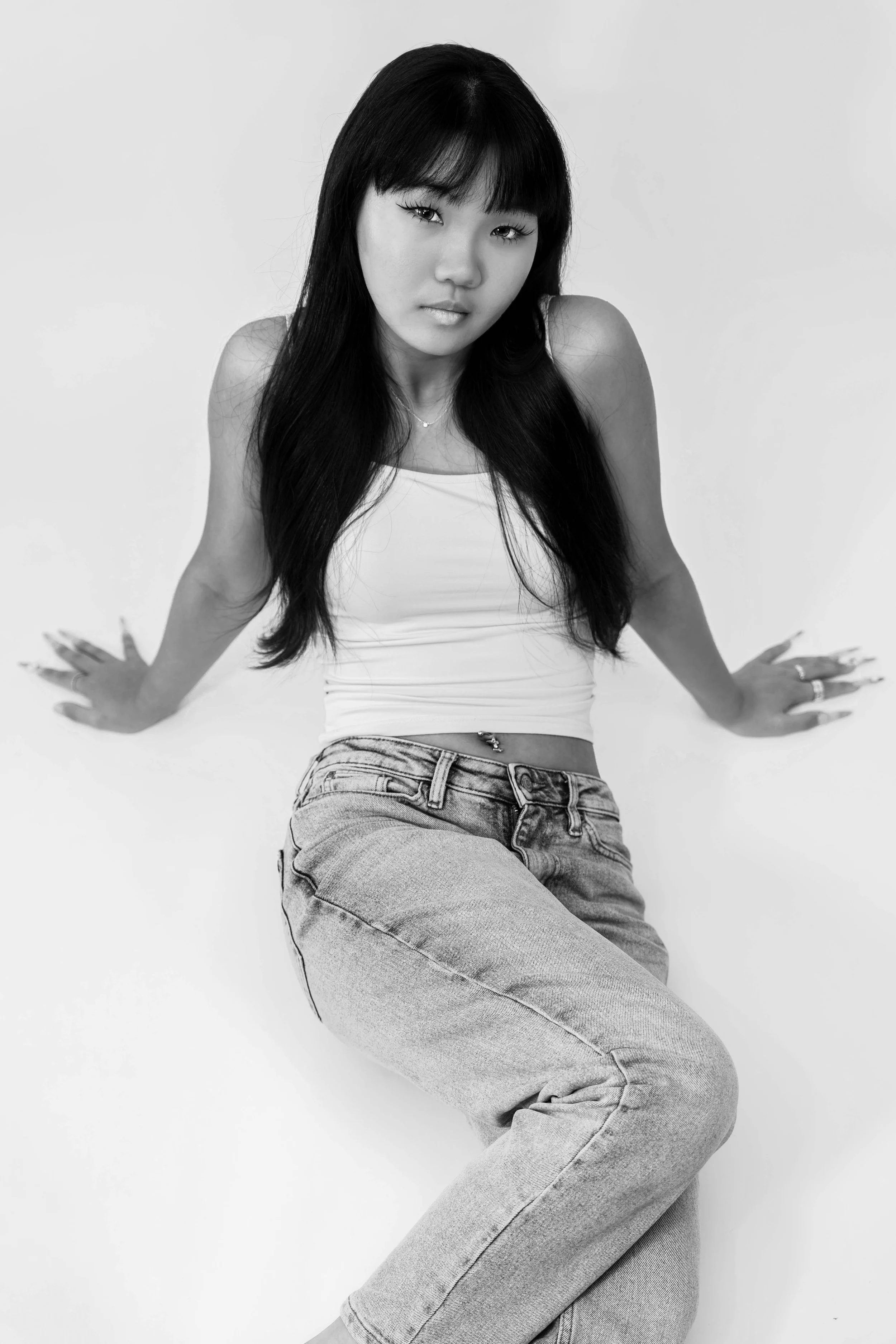 Black and white photograph of a young woman with long dark hair, wearing a tank top and jeans, sitting against a plain background, with a serious expression.